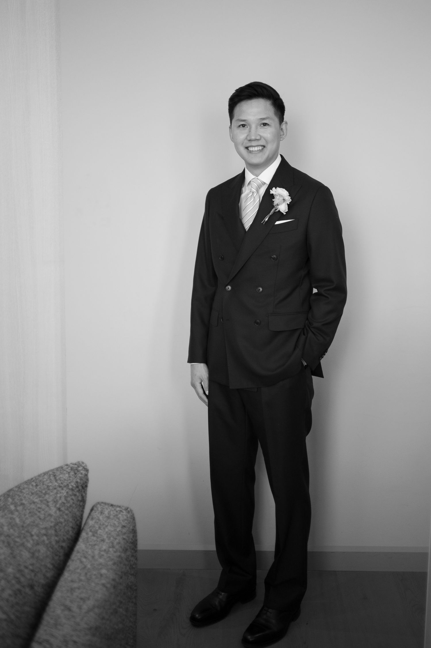 A smiling groom in a black double-breasted suit with boutonniere poses confidently in a black and white wedding photo.