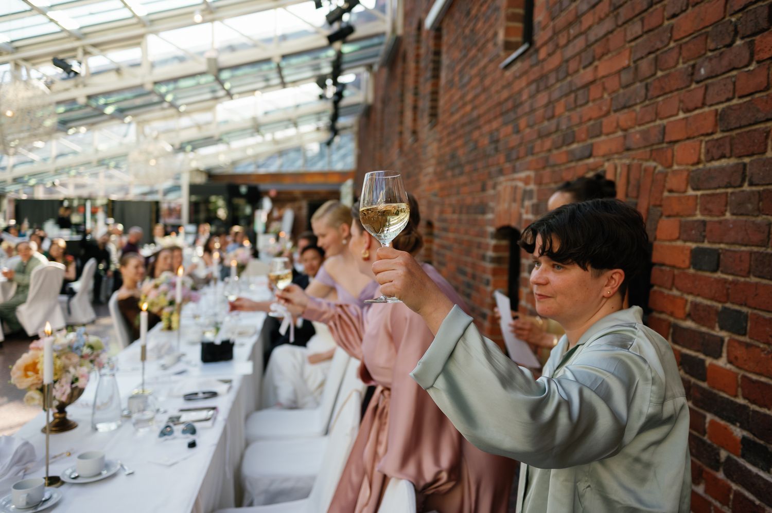 Wedding guests raising glasses of white wine in a toast inside a brick-walled greenhouse reception venue.