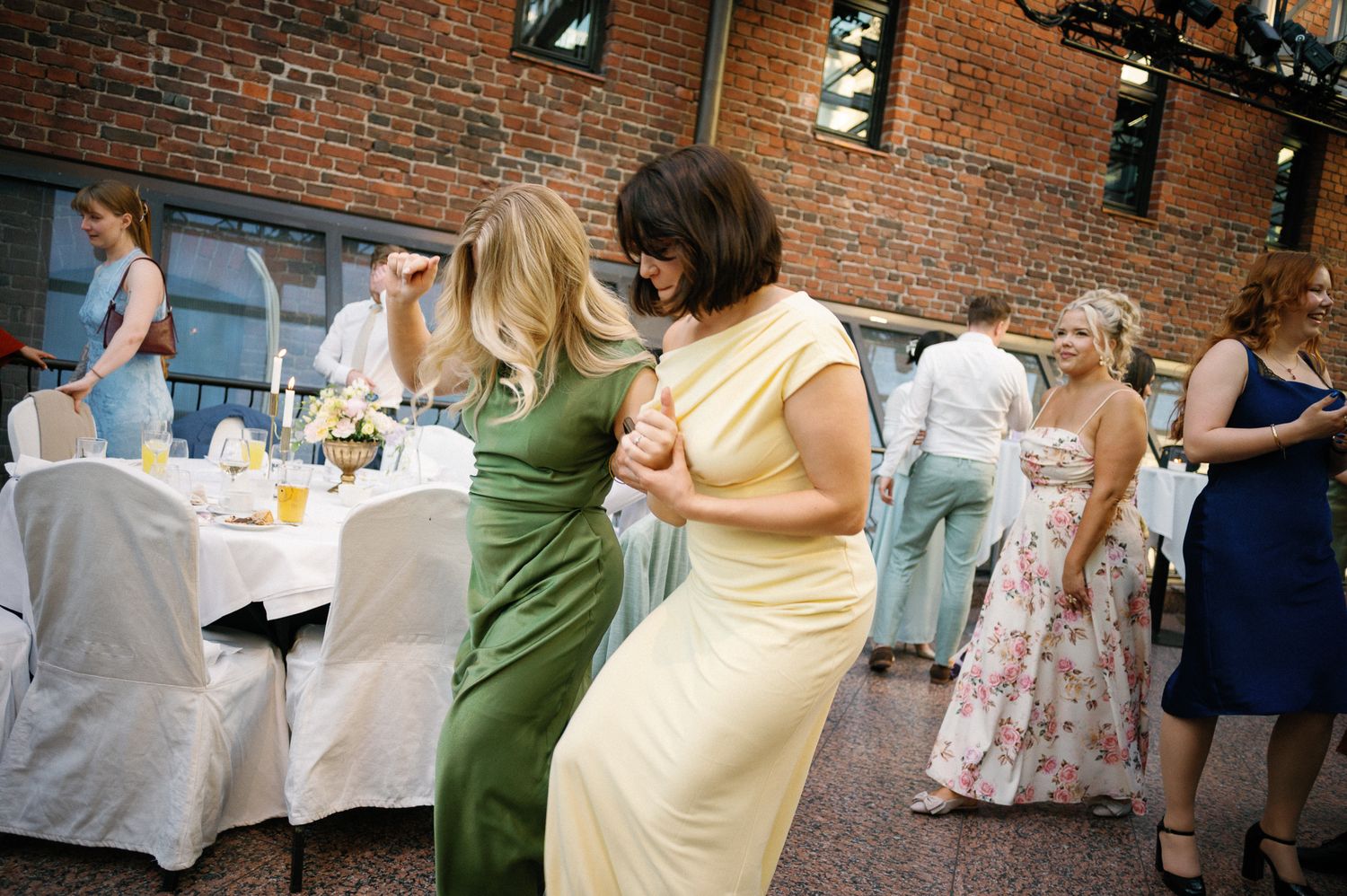 Women in elegant dresses dancing at an outdoor wedding reception with brick wall backdrop.
