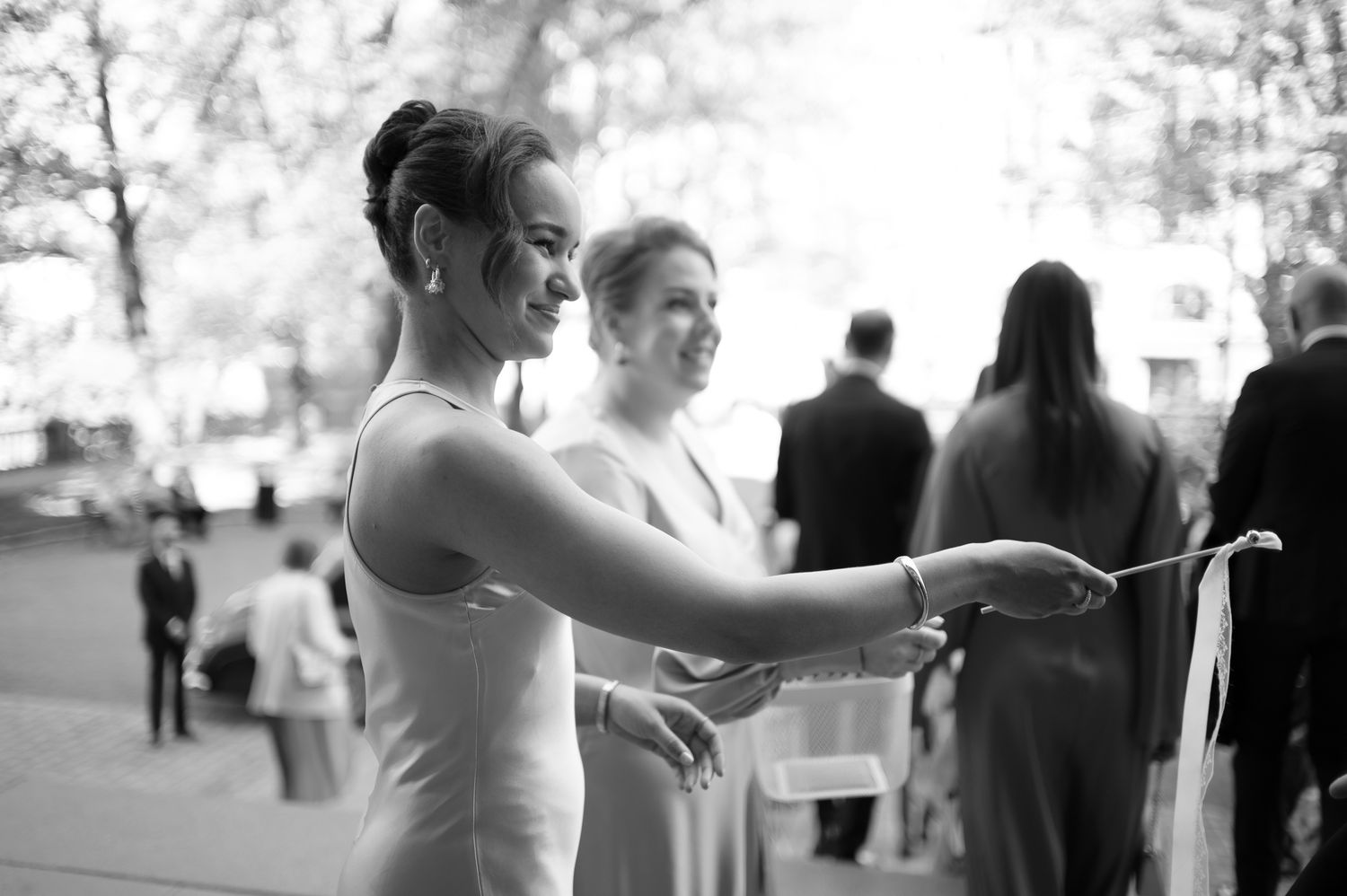 Black and white photo of bride cutting wedding cake outdoors with bridesmaids and guests in background.