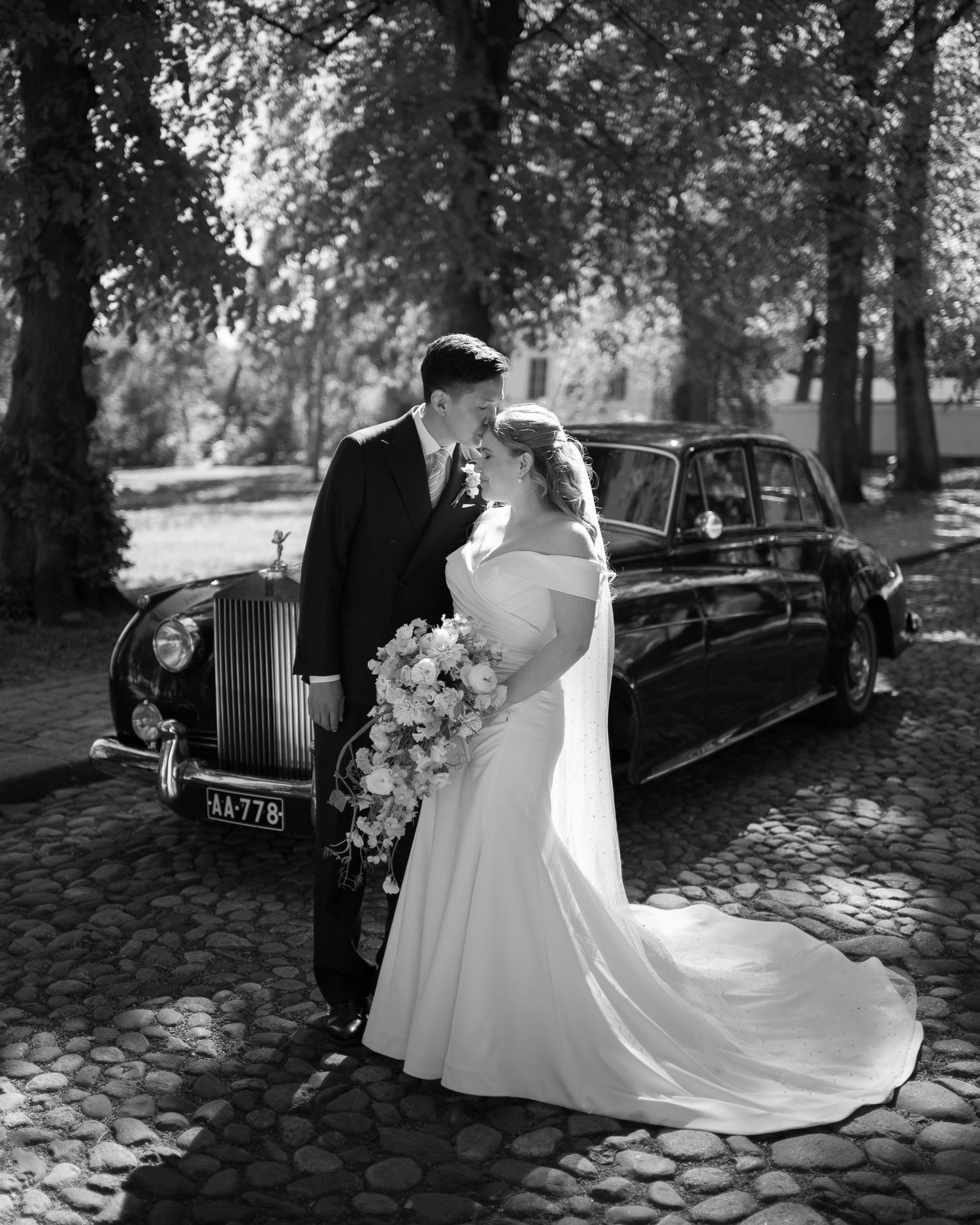 Black and white photo of bride and groom sharing a romantic moment beside a classic Rolls-Royce on cobblestones.