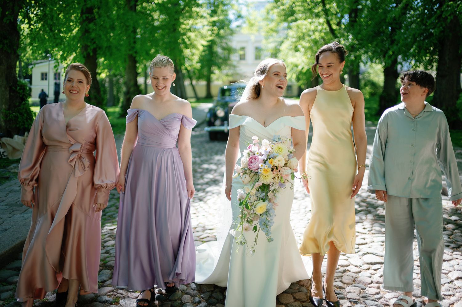 Bride in white gown walks with bridesmaids in pastel dresses on cobblestone path surrounded by lush green trees.
