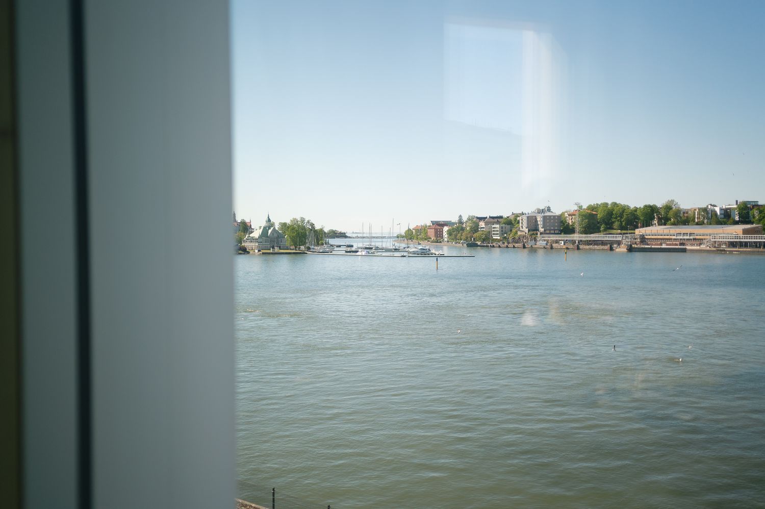 View through a window of a calm river with boats, docks, and tree-lined waterfront buildings under a clear blue sky.