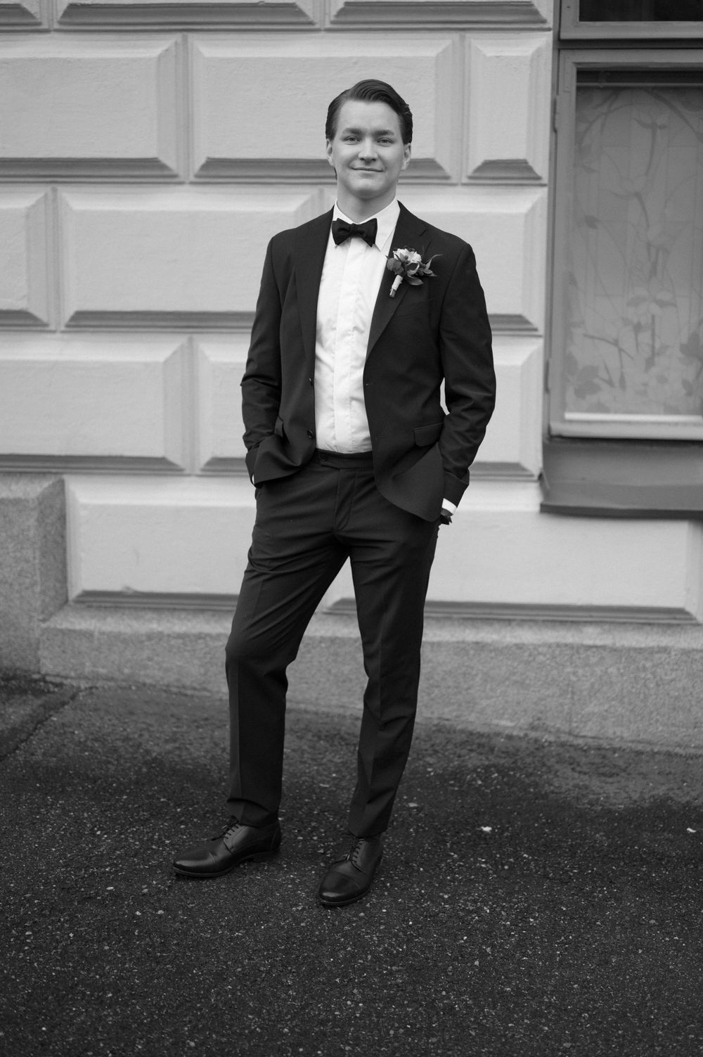 Groom in black tuxedo and bow tie poses casually against light colored wall.