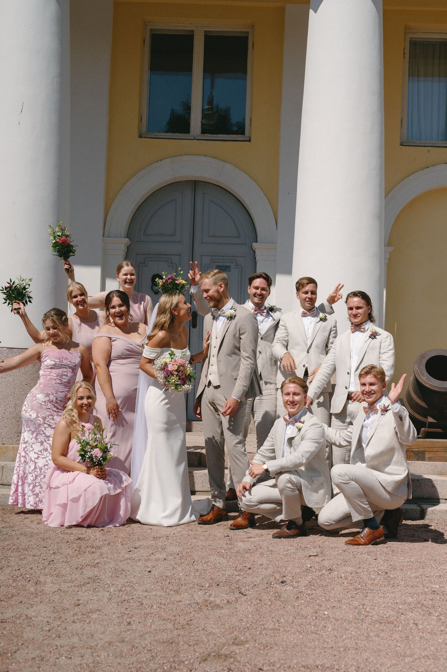 Bridal party in pink dresses and light suits relaxes on stone steps of historic venue.