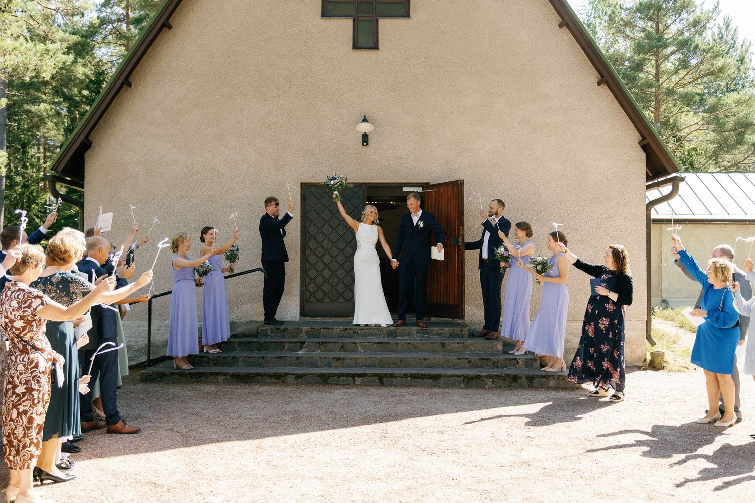 A small white chapel with a cross stands among pine trees during a wedding ceremony.