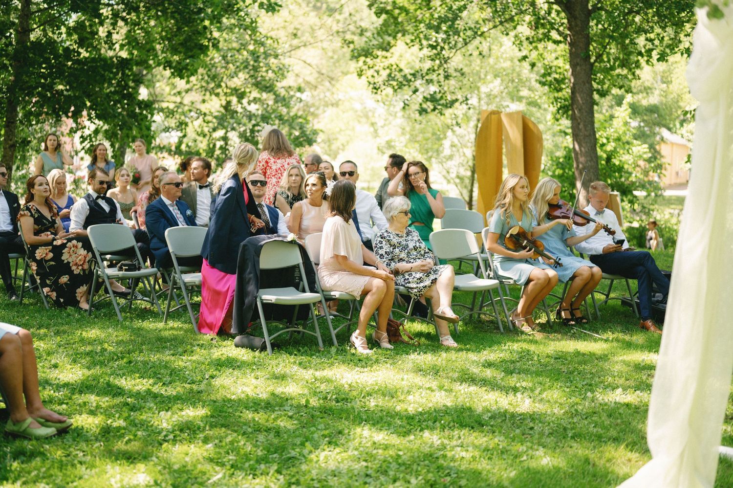 Outdoor wedding reception with guests seated under trees on a sunny day.