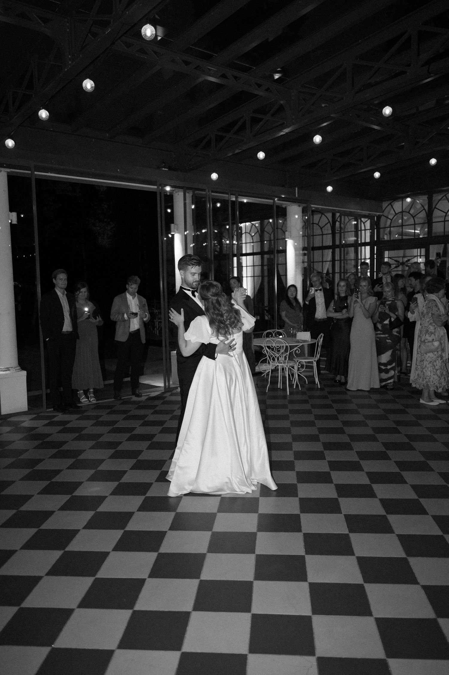 Couples slow dancing on black and white checkered floor under string lights.