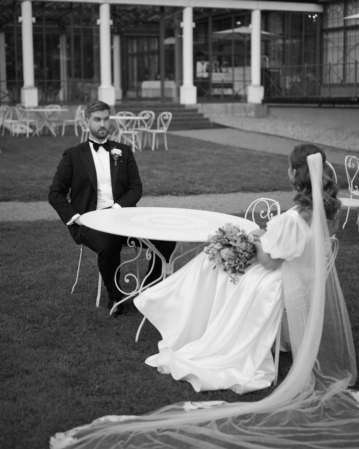 Black and white photo of newlyweds sitting at ornate garden furniture outdoors.