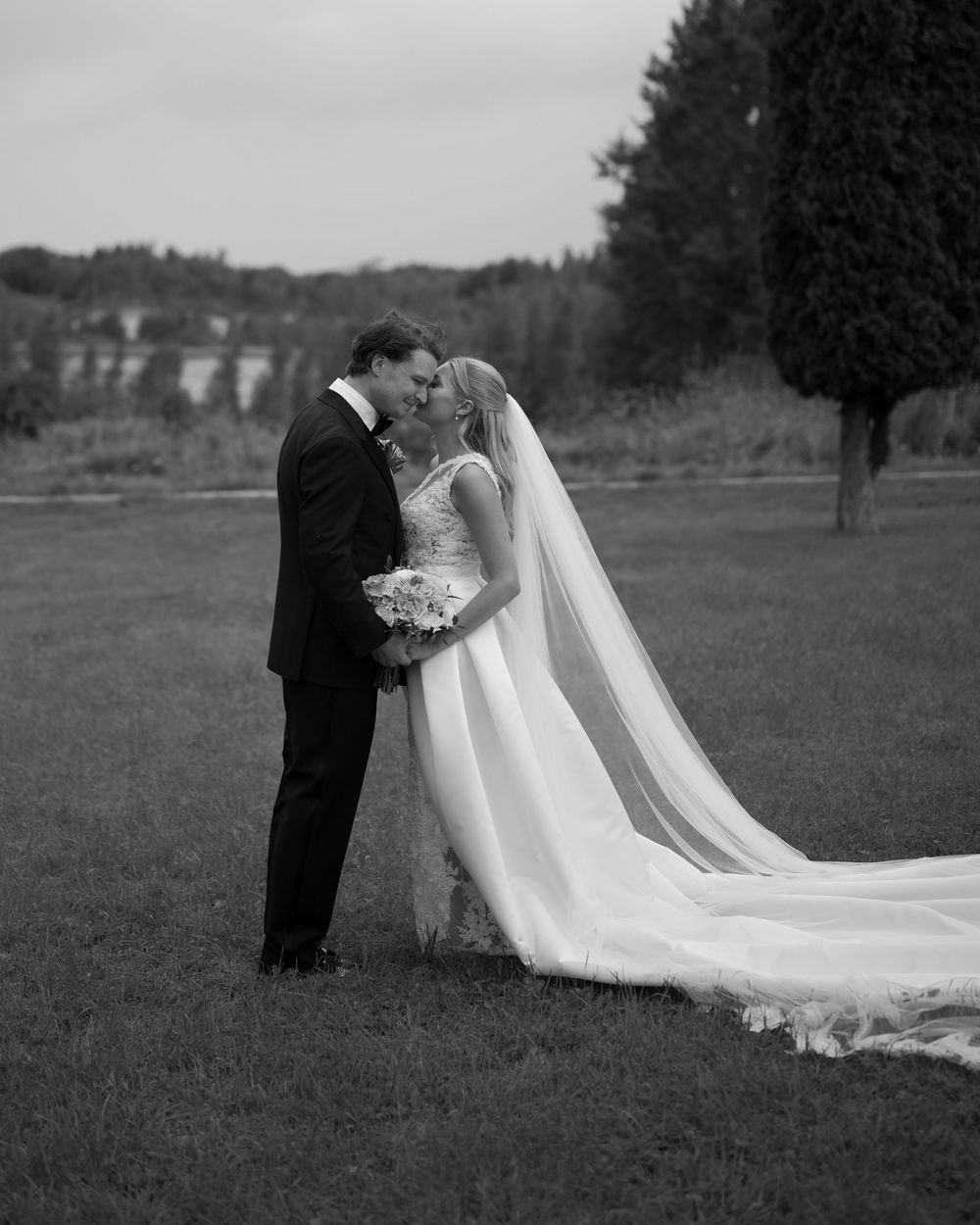 Black and white photo of a bride's flowing train trailing behind on grass.