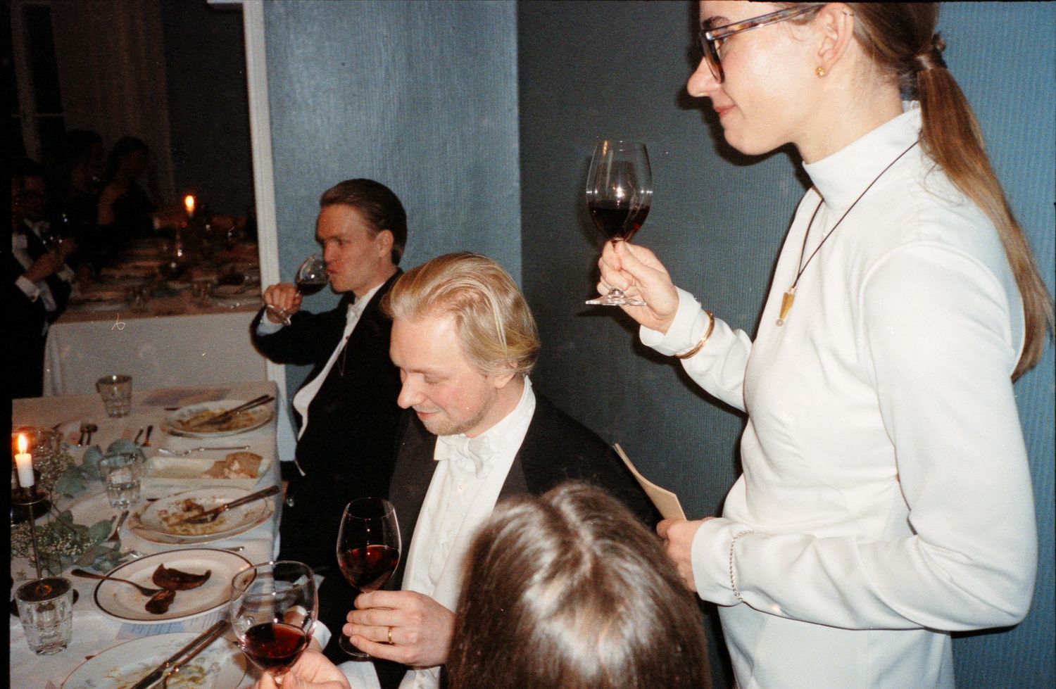 A group of people dining at a formal table setting with wine glasses and white tablecloths in a dimly lit room.