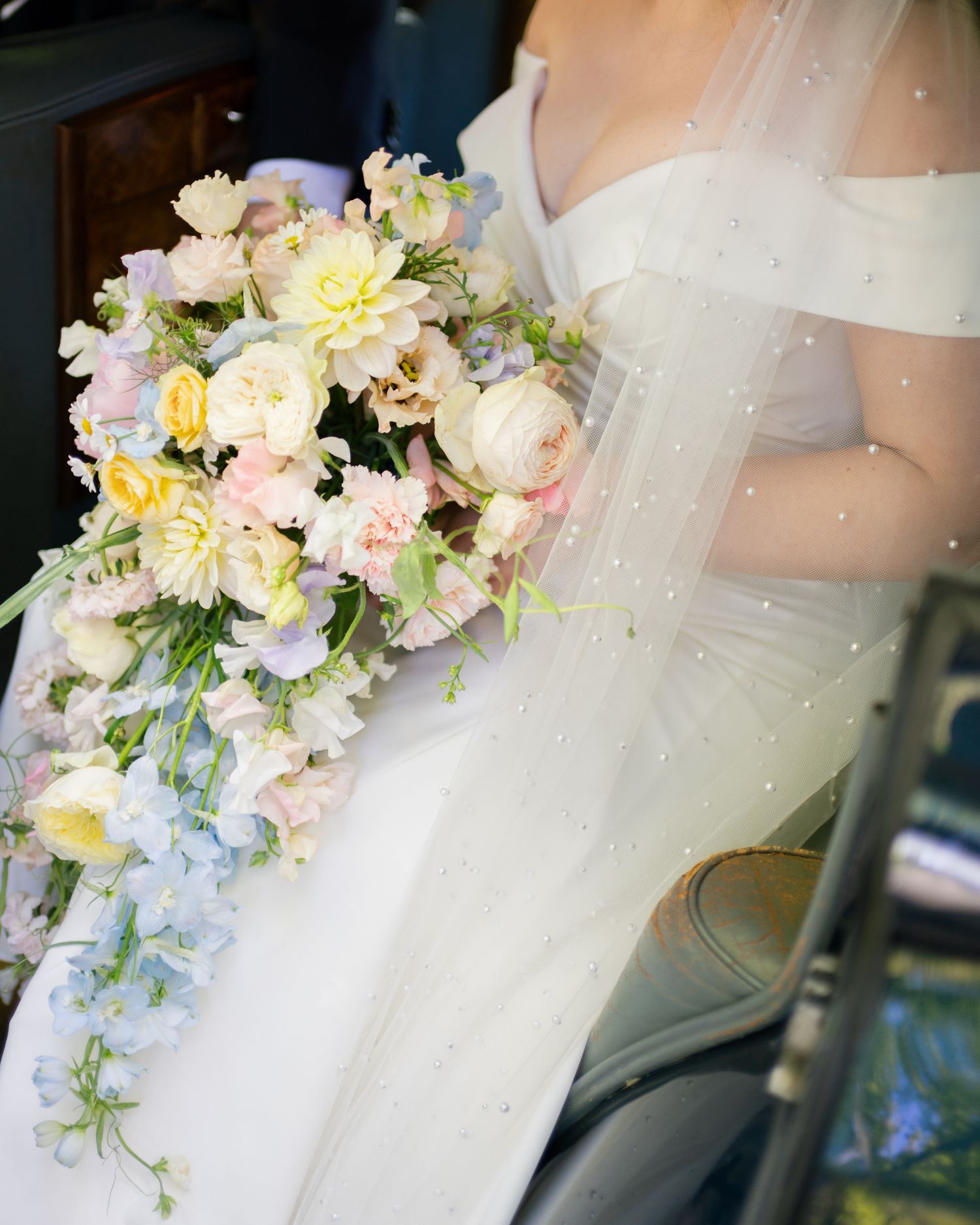 Bride holding a cascading pastel wedding bouquet with pink, yellow, and blue flowers, wearing a white gown with veil.