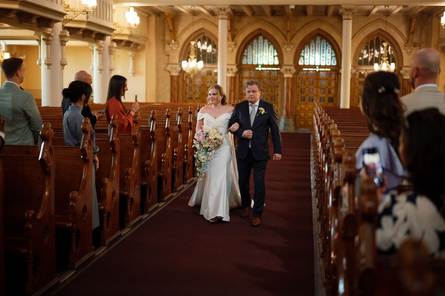 Bride walks down the aisle with escort in elegant church wedding ceremony with wooden pews and ornate architecture.