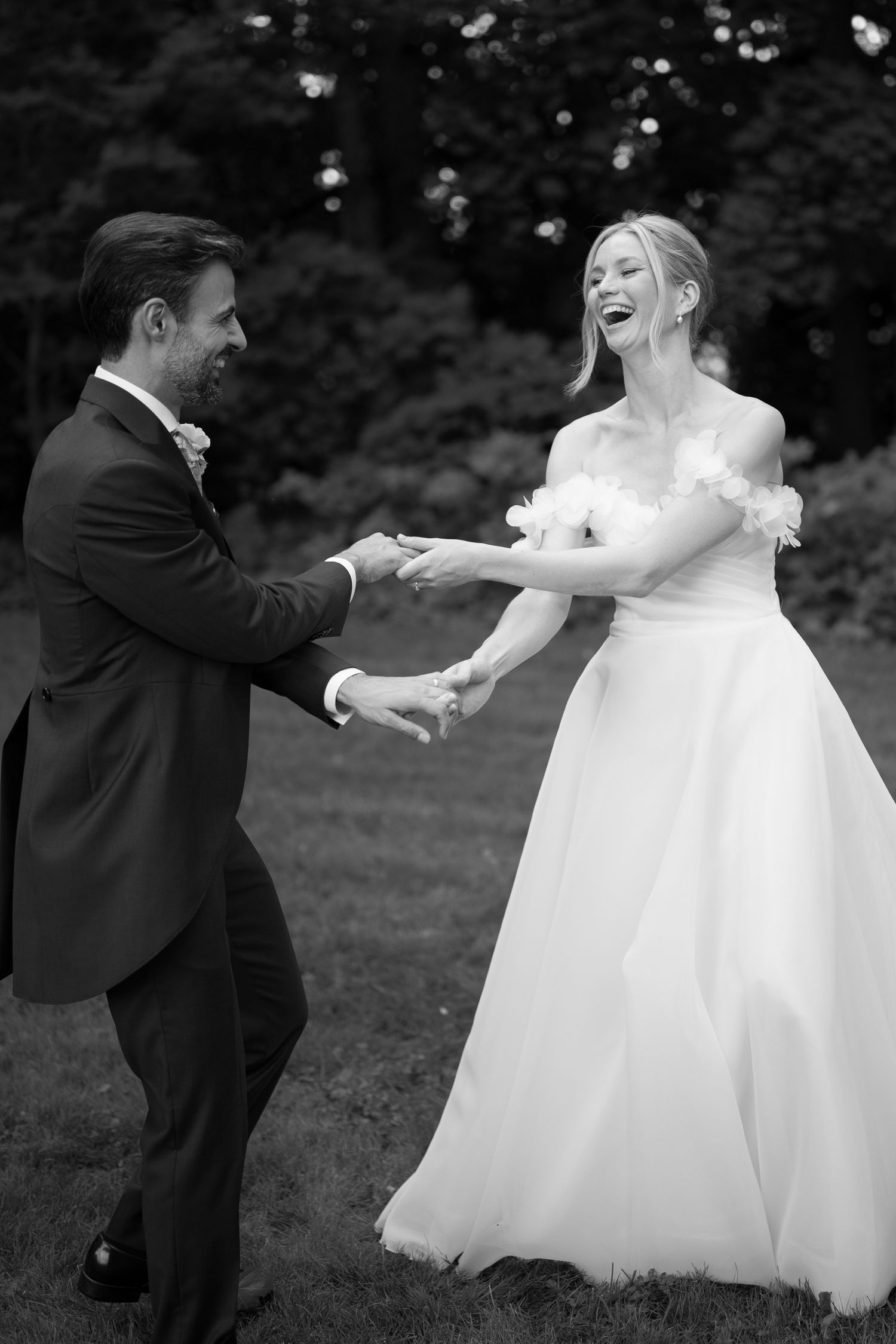 Black and white photo of a laughing bride in a white gown dancing with a groom in a dark suit outdoors.