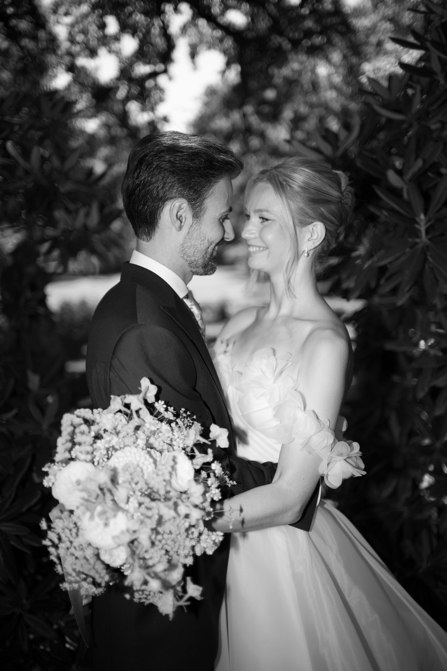 Black and white wedding photo of bride and groom gazing at each other, bride holding floral bouquet, outdoors.
