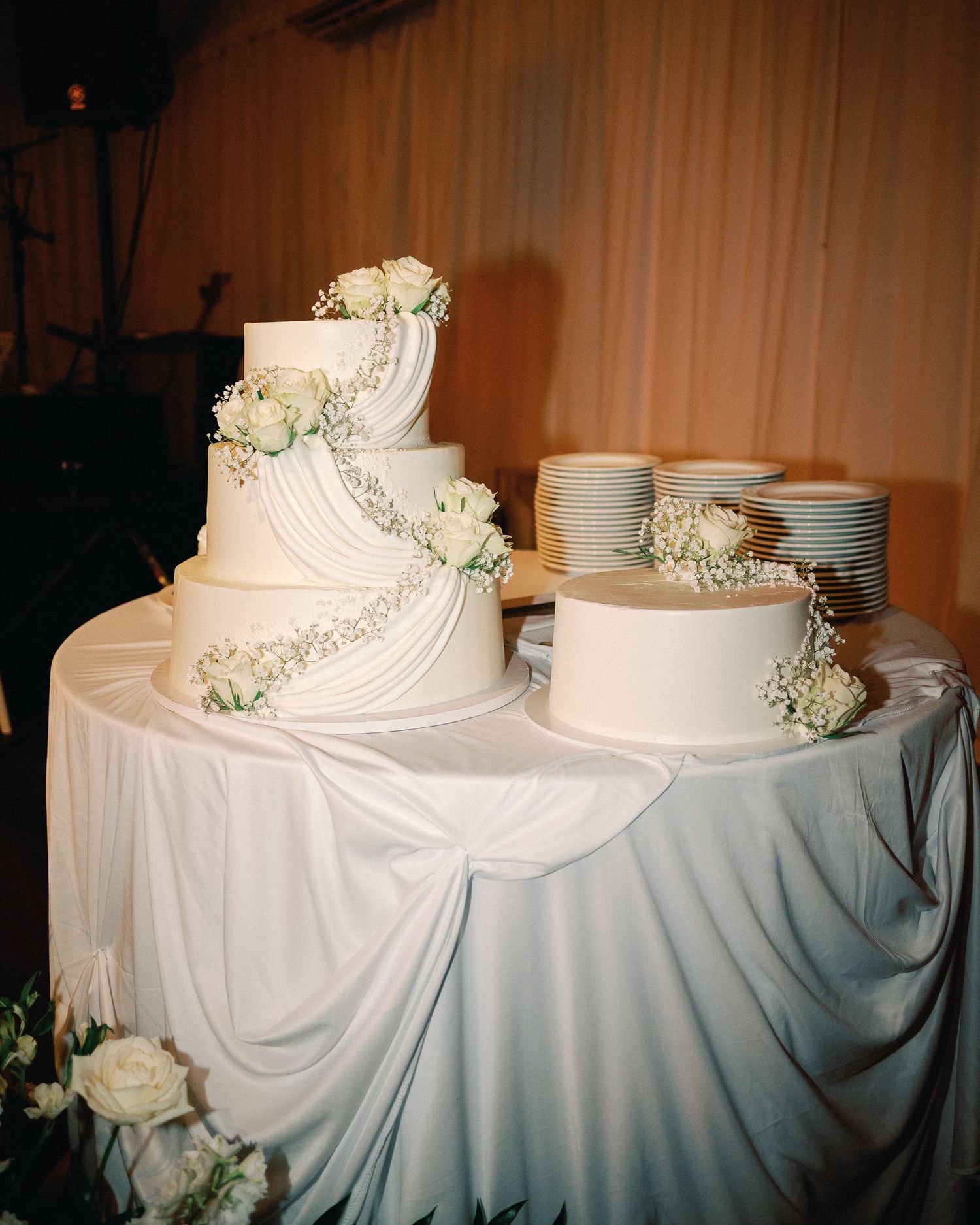 Elegant three-tier white wedding cake adorned with roses and baby's breath on a draped satin table.