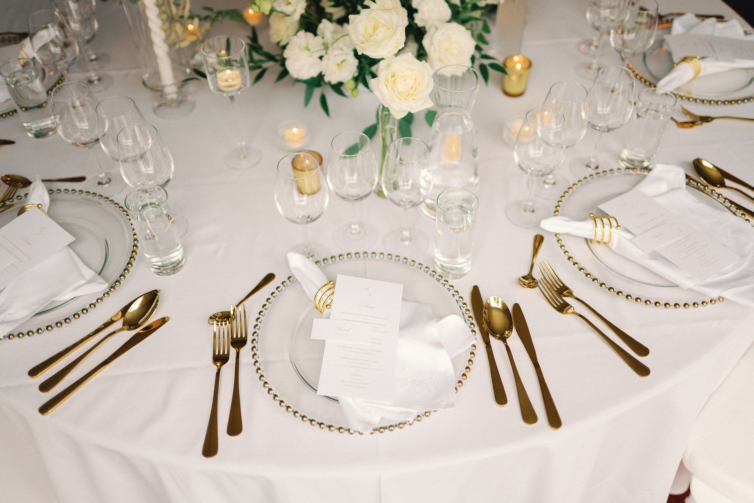Elegant wedding reception table setting with gold cutlery, pearl-edged charger plates, crystal glasses, and white rose centerpiece.