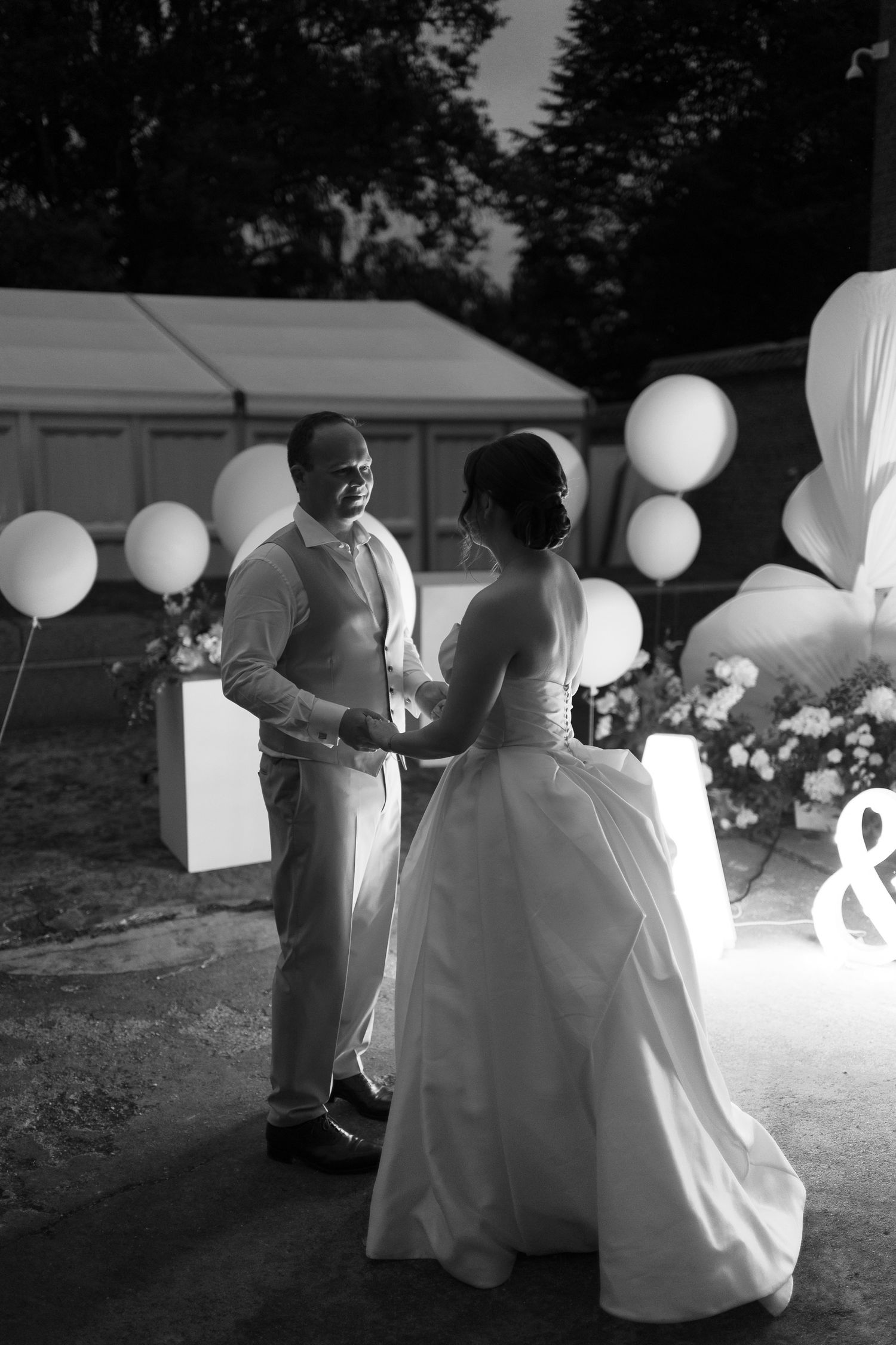 Black and white photo of bride and groom holding hands outdoors at night with balloons and floral decorations.