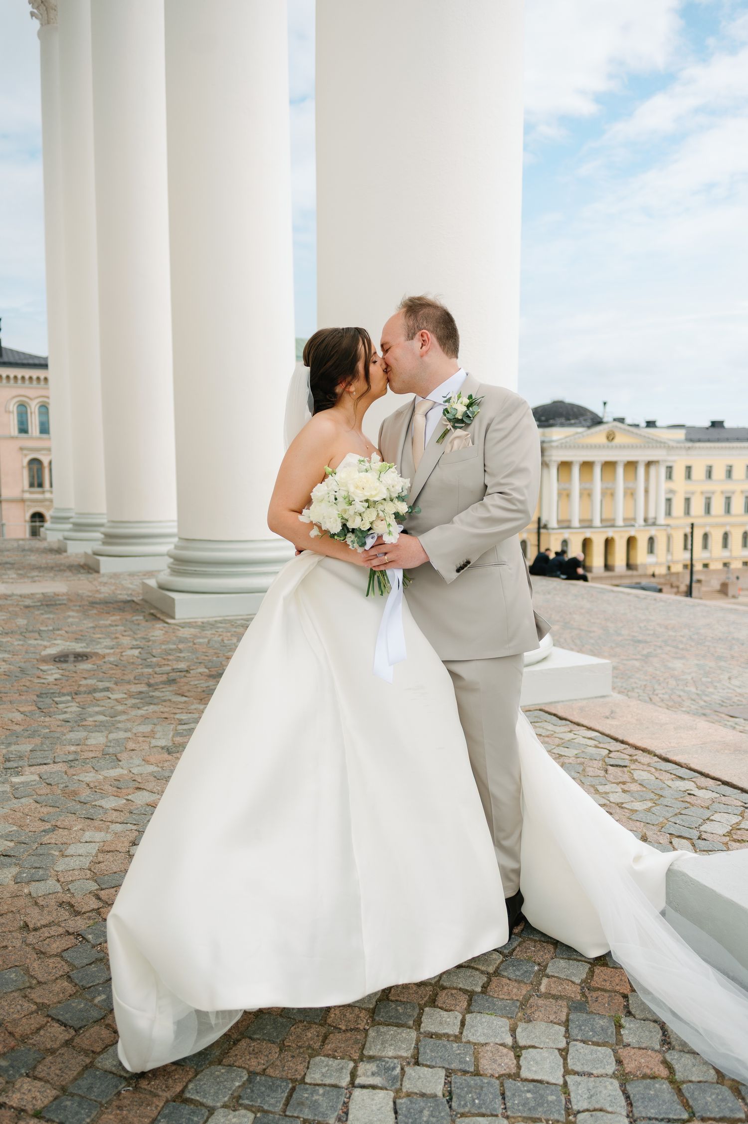 Bride and groom share a romantic kiss near white columns, bride holding white bouquet in elegant strapless gown, groom in tan suit.