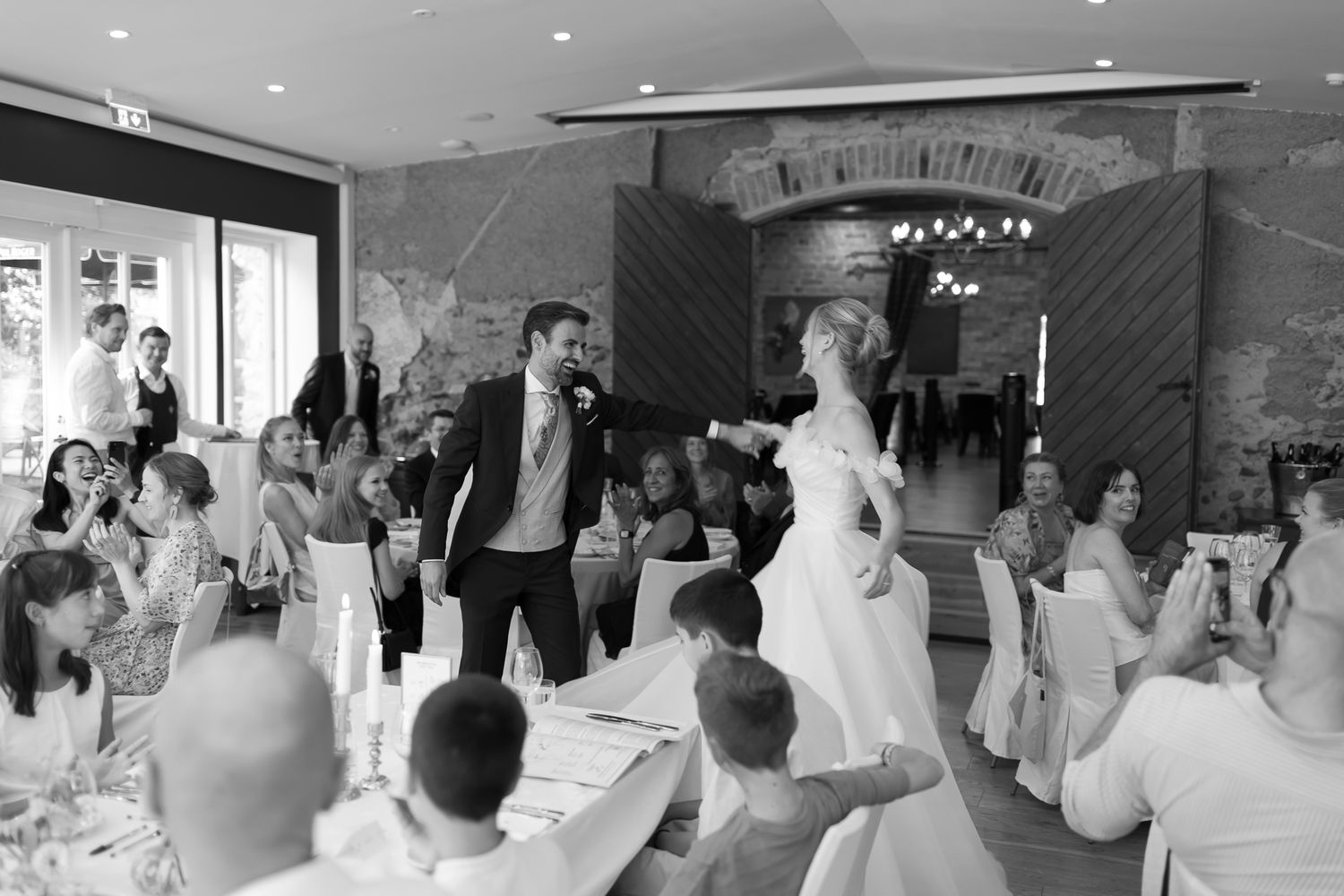 Black and white wedding photo of bride and groom entering reception hall, guests seated at decorated tables.