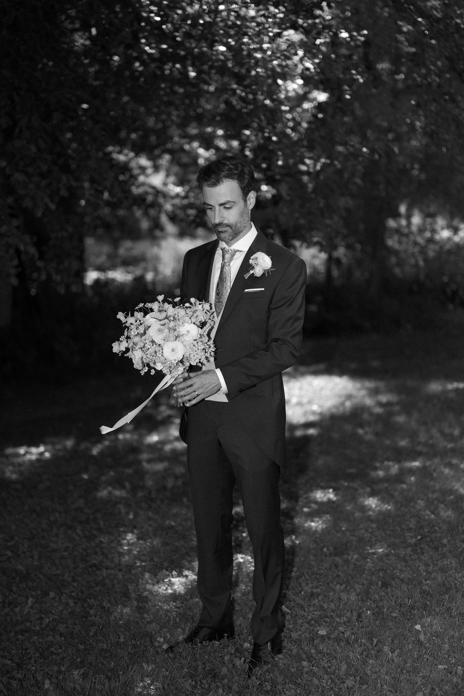 Black and white photo of a groom in a suit holding a floral bouquet outdoors among trees.