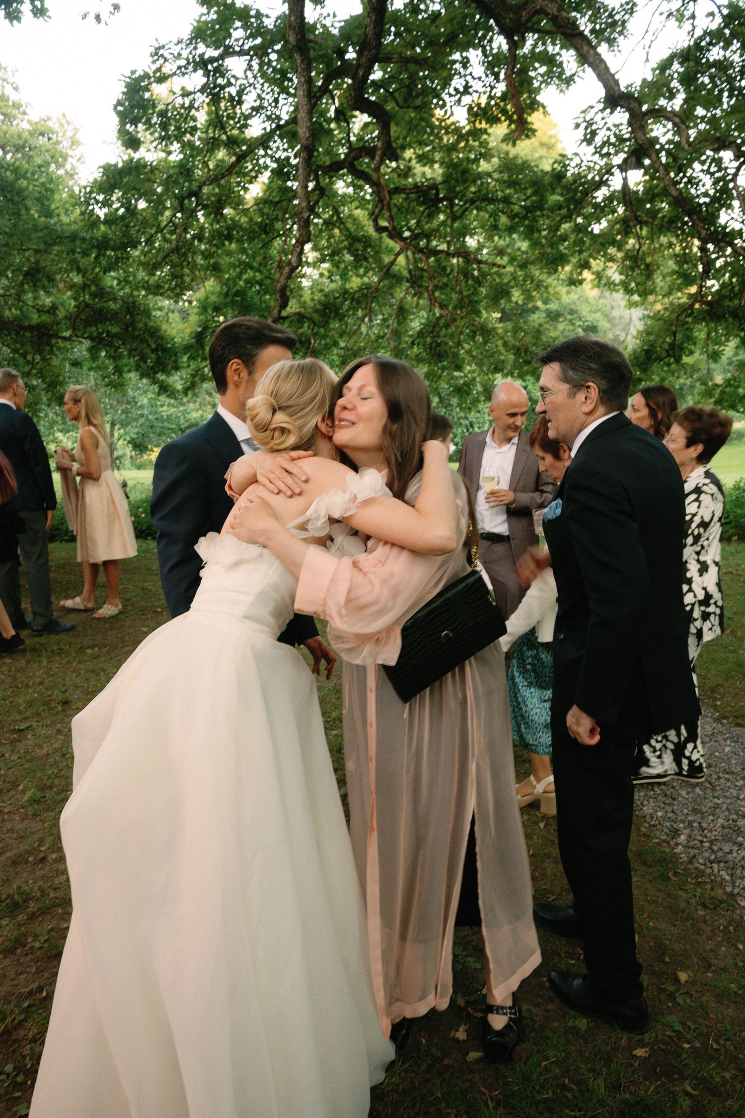 Bride and groom sharing warm hugs with wedding guests outdoors under lush green trees at a garden reception.