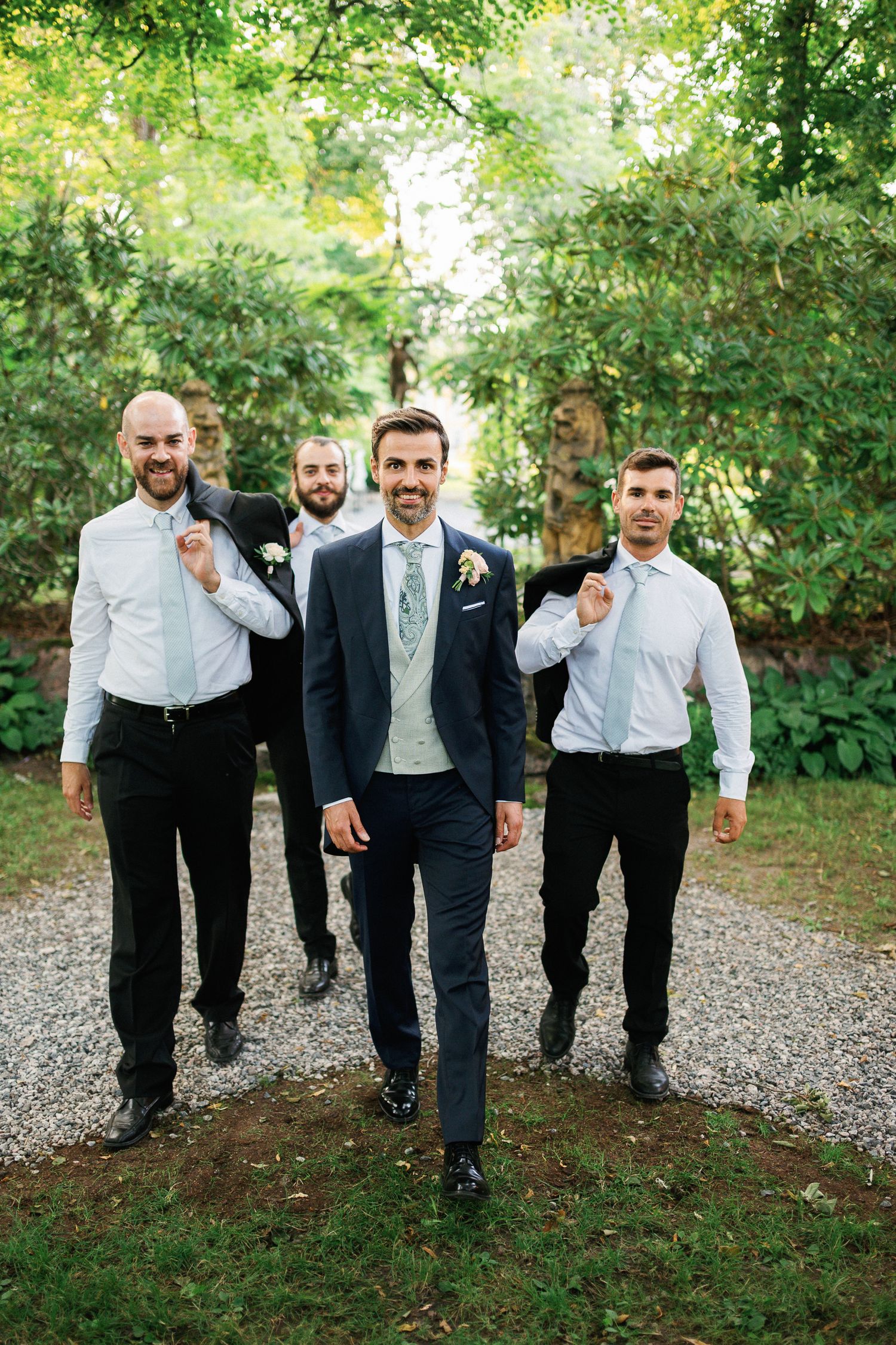 Groom in navy suit leads three groomsmen in white shirts walking on gravel path surrounded by lush green trees.