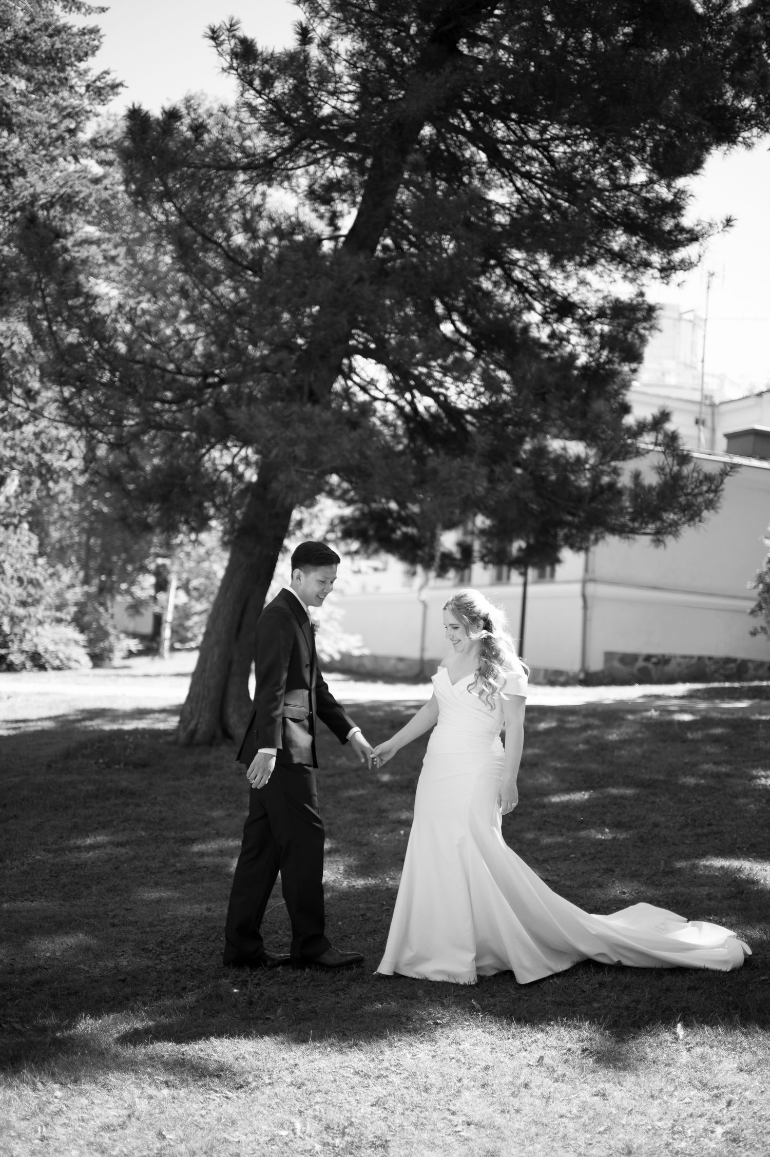 Black and white photo of a bride and groom holding hands in a sunlit park with large trees.