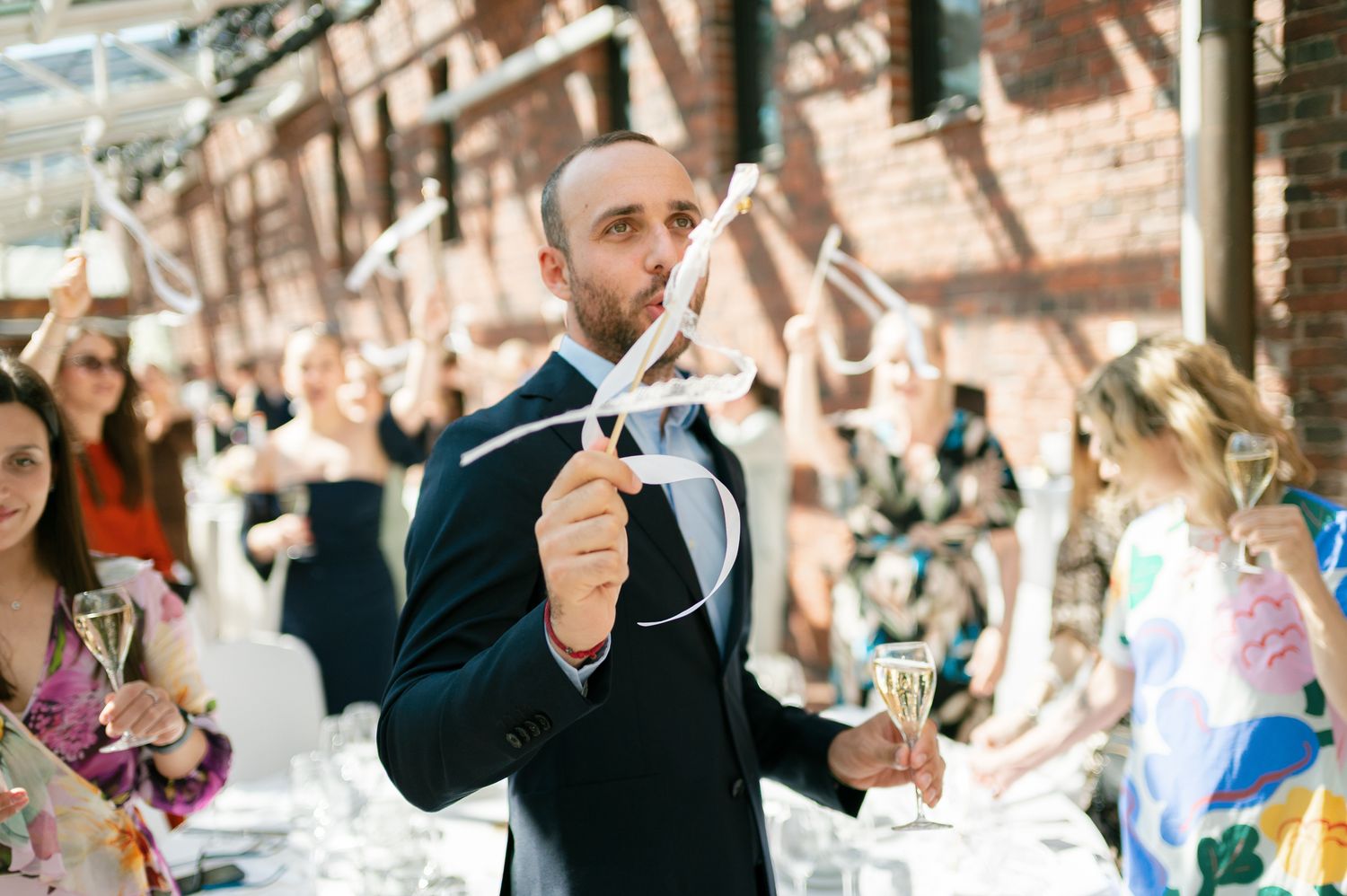 A man in a navy suit holds a ribbon wand and champagne glass at an outdoor wedding celebration.