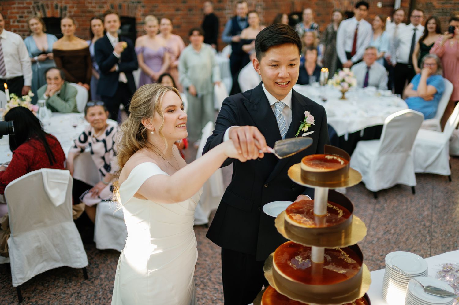 Bride and groom cutting a tiered wedding cake together while guests celebrate in the background.