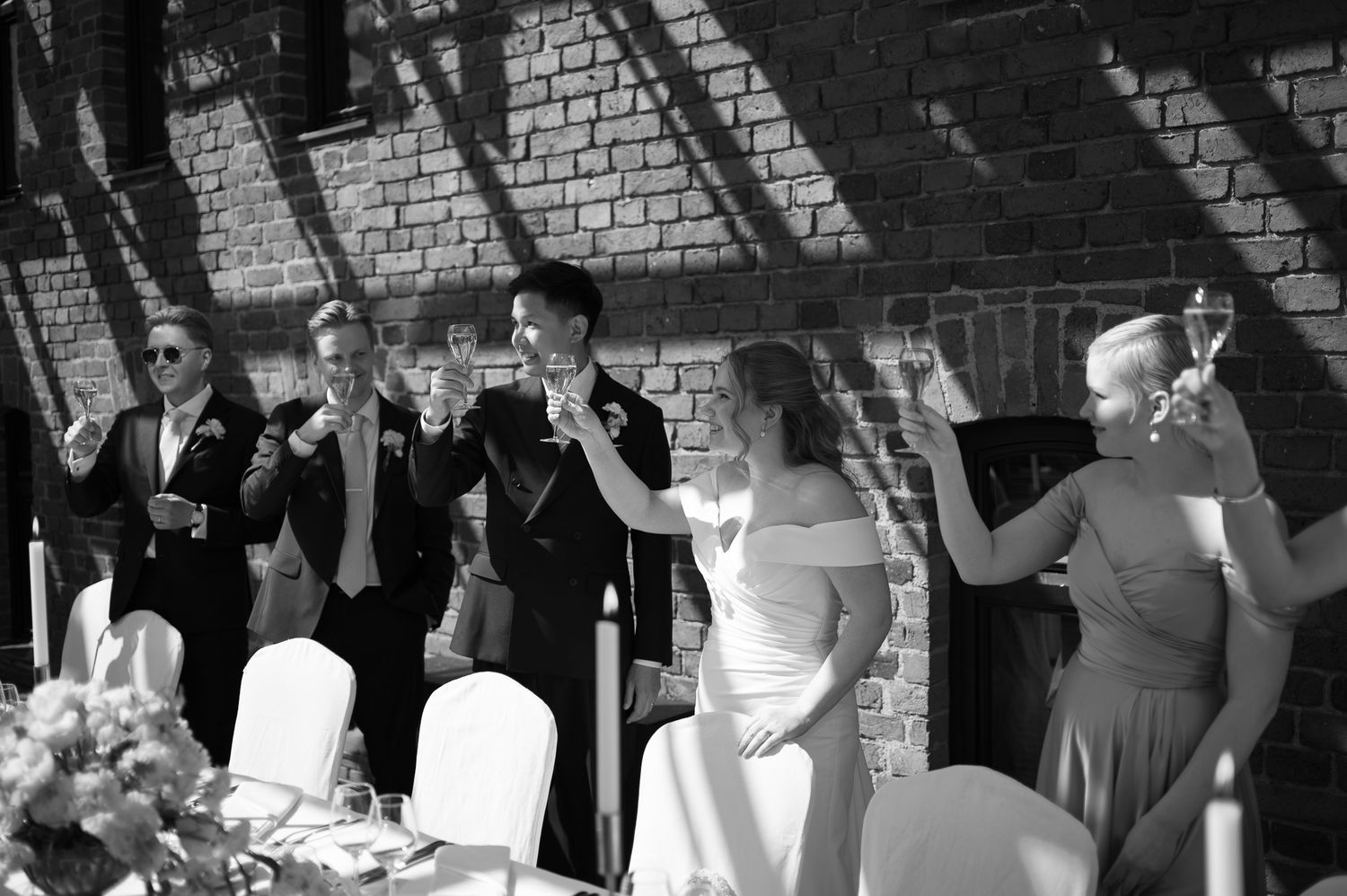 Black and white wedding toast with bride, groom, and guests raising glasses near brick wall.