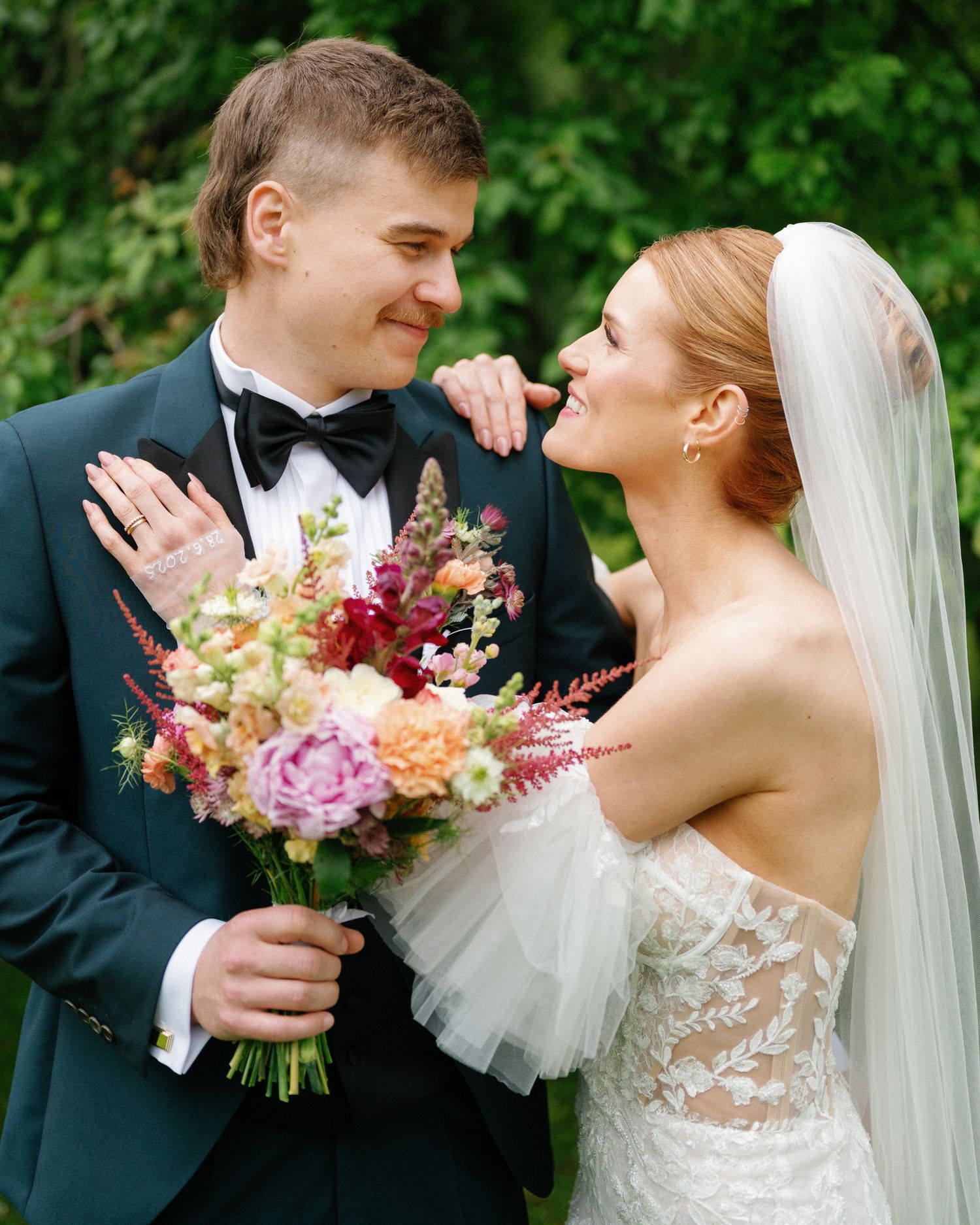Bride and groom share a romantic moment, she holds a colorful bouquet, he wears a dark tuxedo, outdoors.