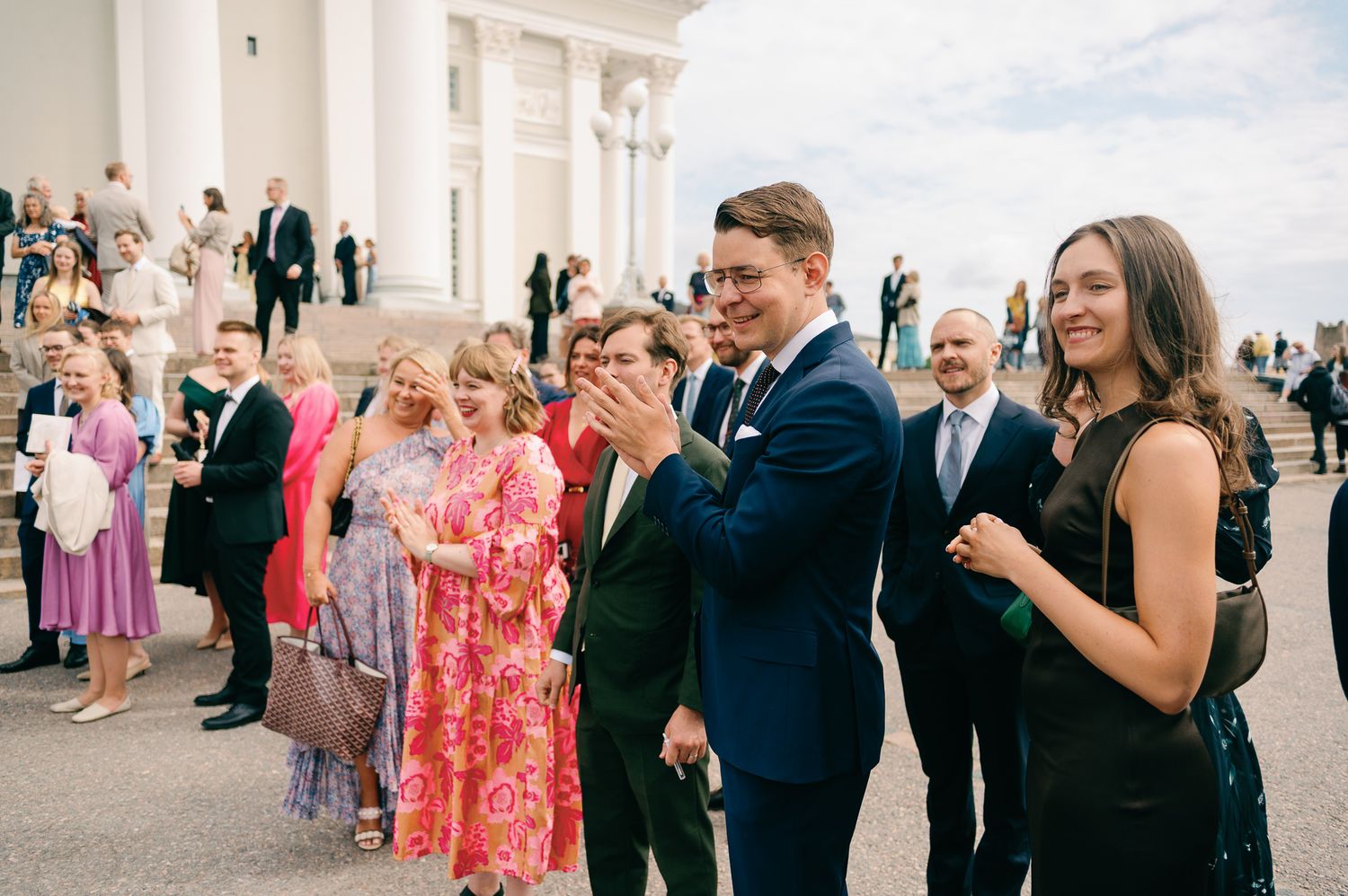 Wedding guests clapping and smiling on church steps, dressed in formal attire at an outdoor ceremony.