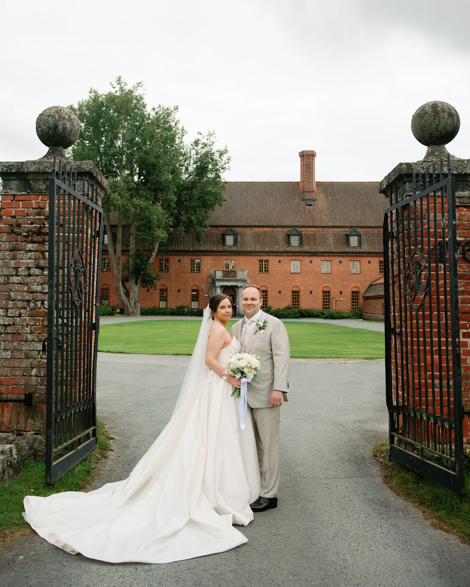 Bride and groom pose at ornate iron gates of historic red brick estate on their wedding day.
