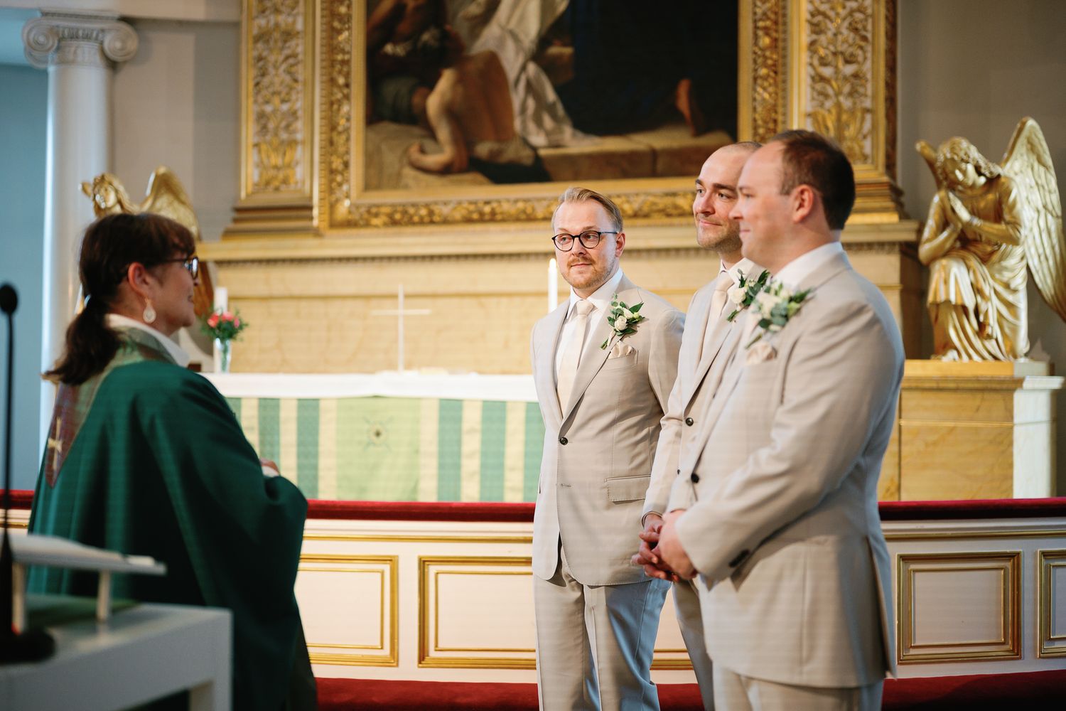 Two grooms in beige suits exchange vows with an officiant at a church altar during a same-sex wedding ceremony.