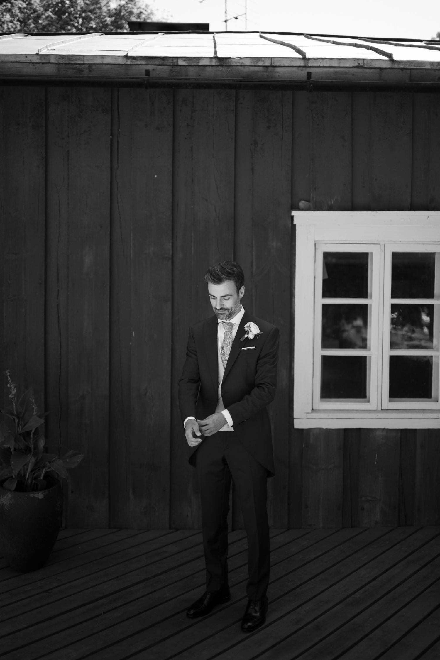 Black and white photo of a groom in a suit adjusting his cufflinks on a wooden deck outside a rustic barn.