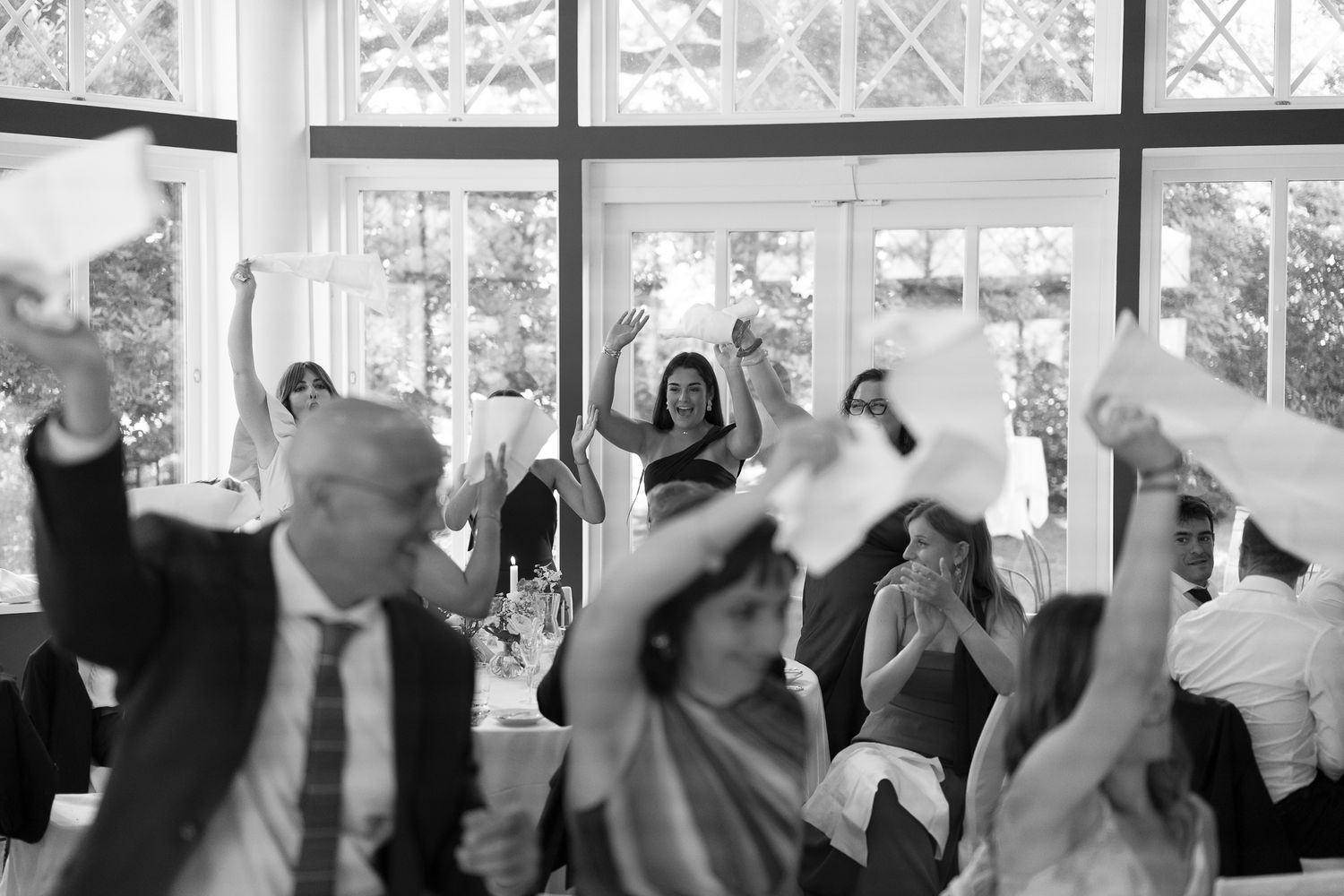 Black and white wedding reception photo of guests joyfully waving napkins in the air inside an elegant venue.