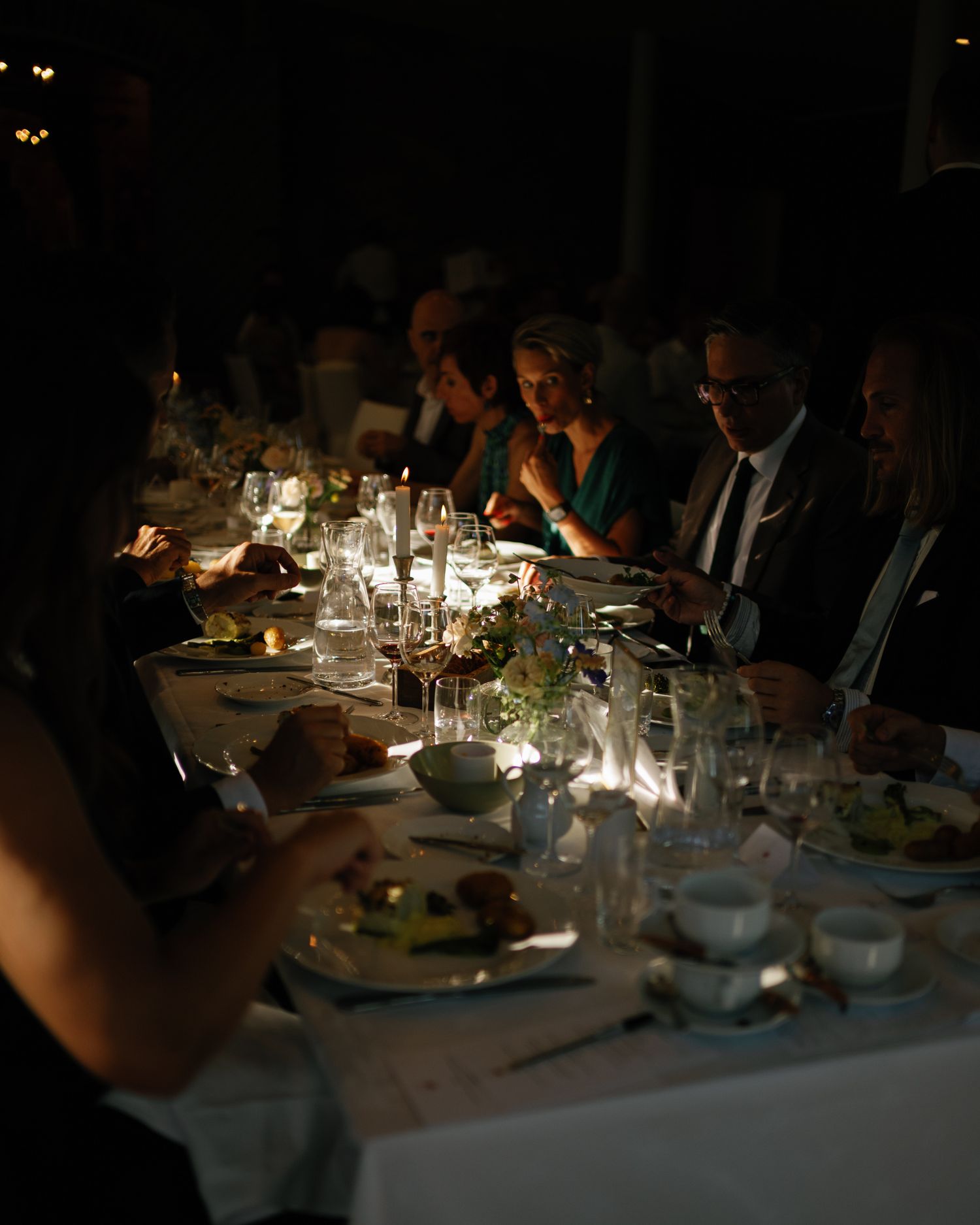 Elegant candlelit dinner party with guests seated at a long table set with glassware and floral centerpieces in dim lighting.
