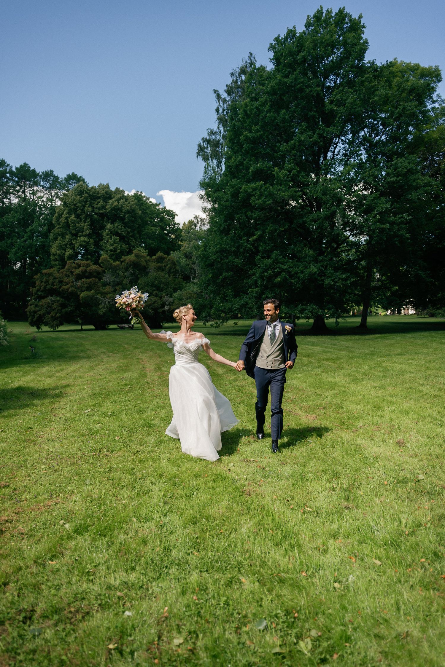 Bride and groom holding hands running joyfully across green lawn with bouquet, trees and blue sky backdrop.