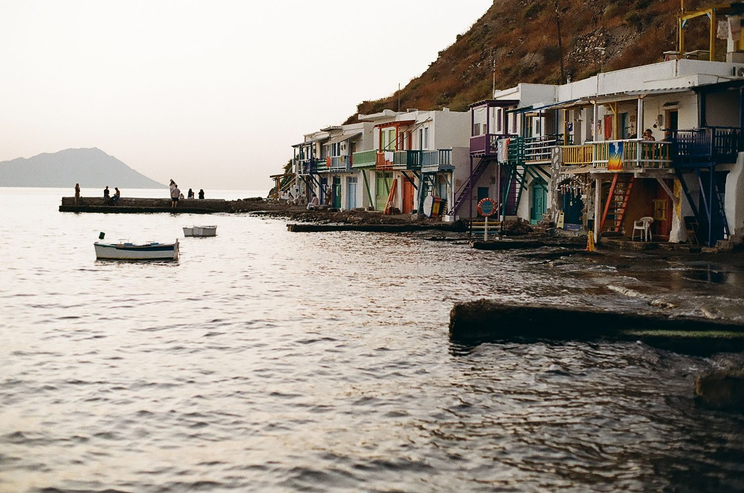 Colorful waterfront houses on stilts line the calm sea in Klima village, Milos, Greece, with small boats floating nearby.