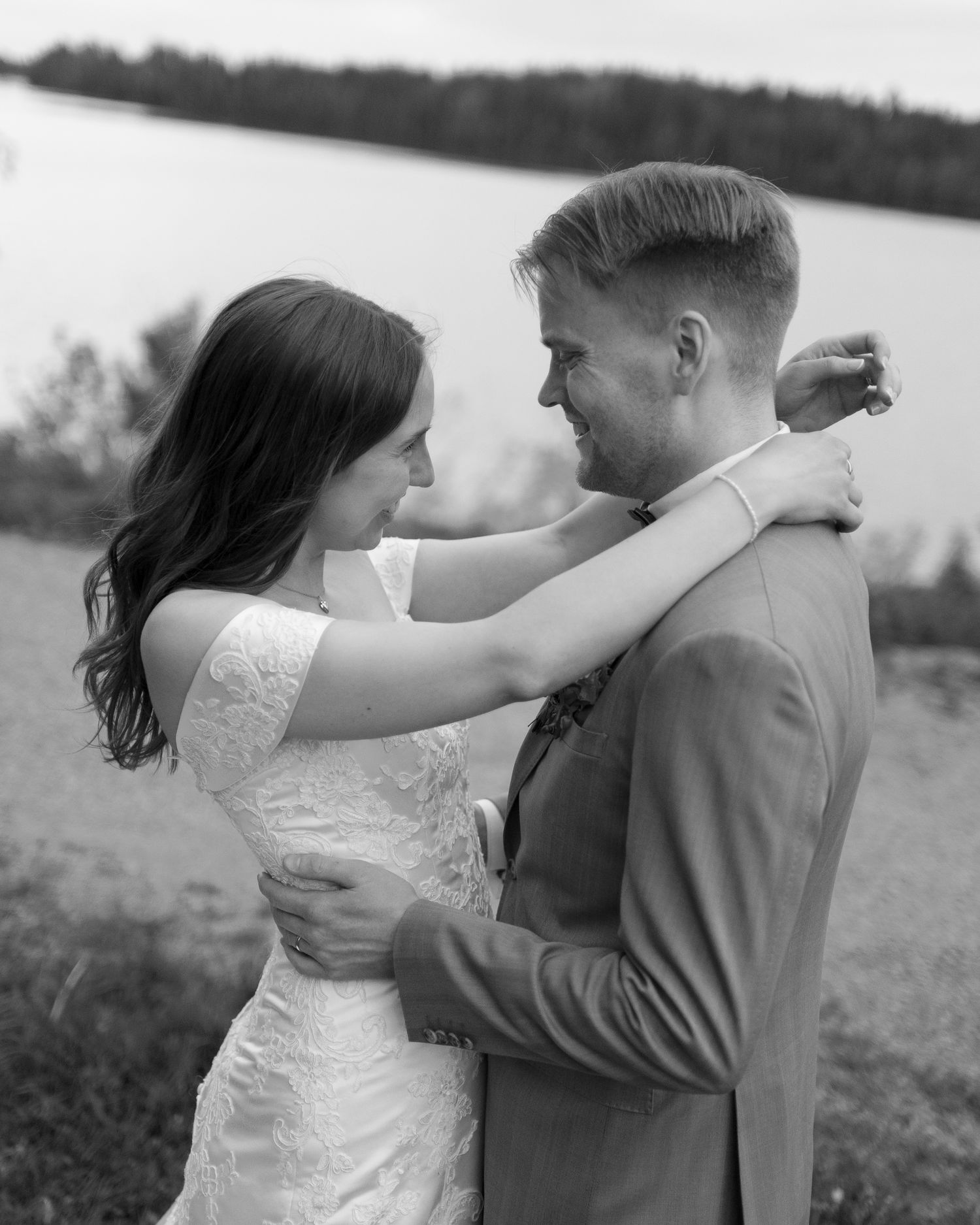Black and white photo of a bride and groom embracing near a lake, sharing a tender moment on their wedding day.