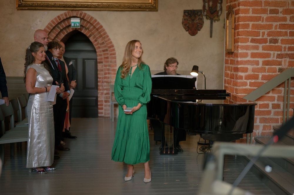 A woman in an emerald green dress stands near a grand piano during a wedding ceremony.