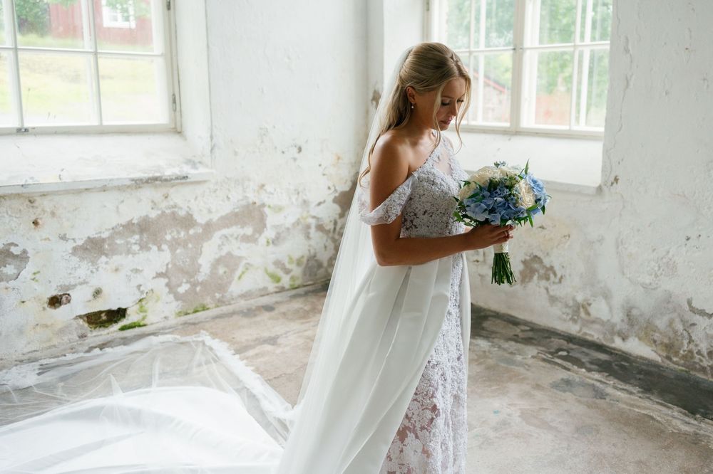 A bride in a lace gown poses in a bright white room with distressed walls and large windows.