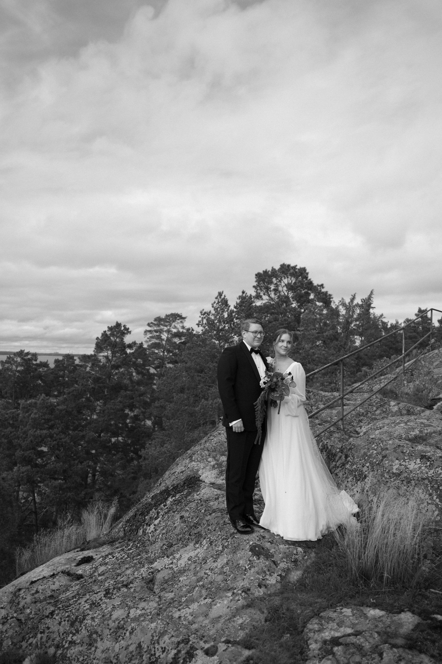 Black and white wedding photograph of a couple standing together on a hilltop with trees and cloudy sky in the background.
