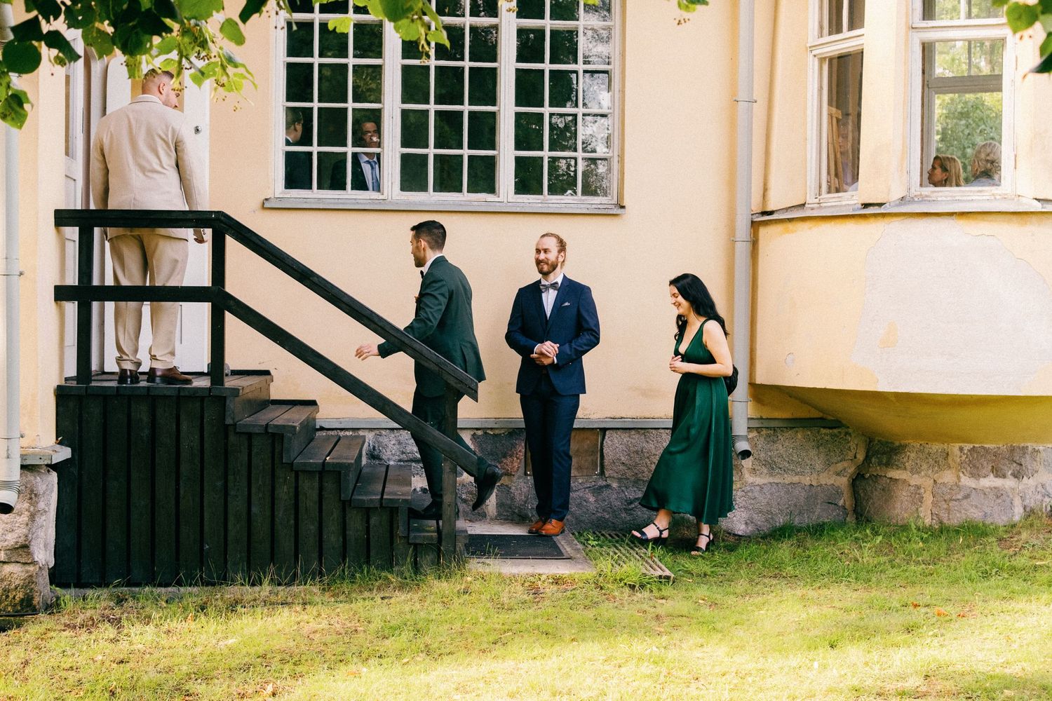 Guests in formal attire gathering on the steps of a yellow building with large windows.