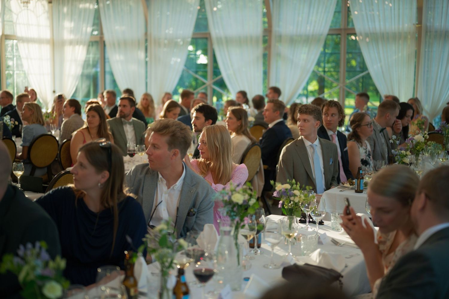 Wedding guests seated at decorated tables in a bright reception hall with natural lighting.