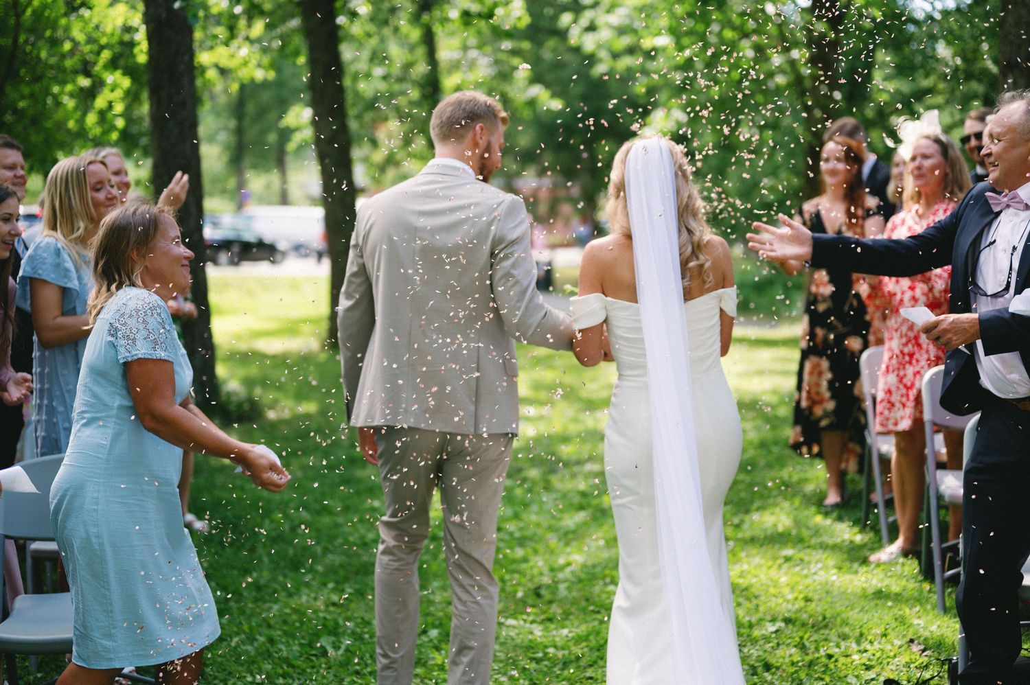 Wedding guests watch the ceremony from white chairs arranged on a grassy lawn under tall trees.