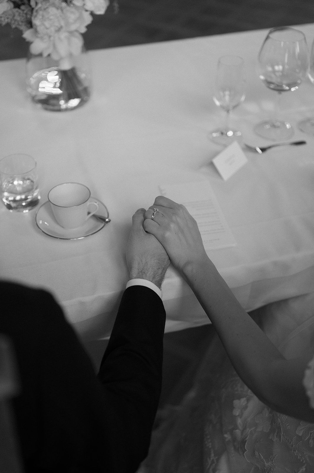 Black and white image of hands joined across a formal dining table with teacups and wine glasses.