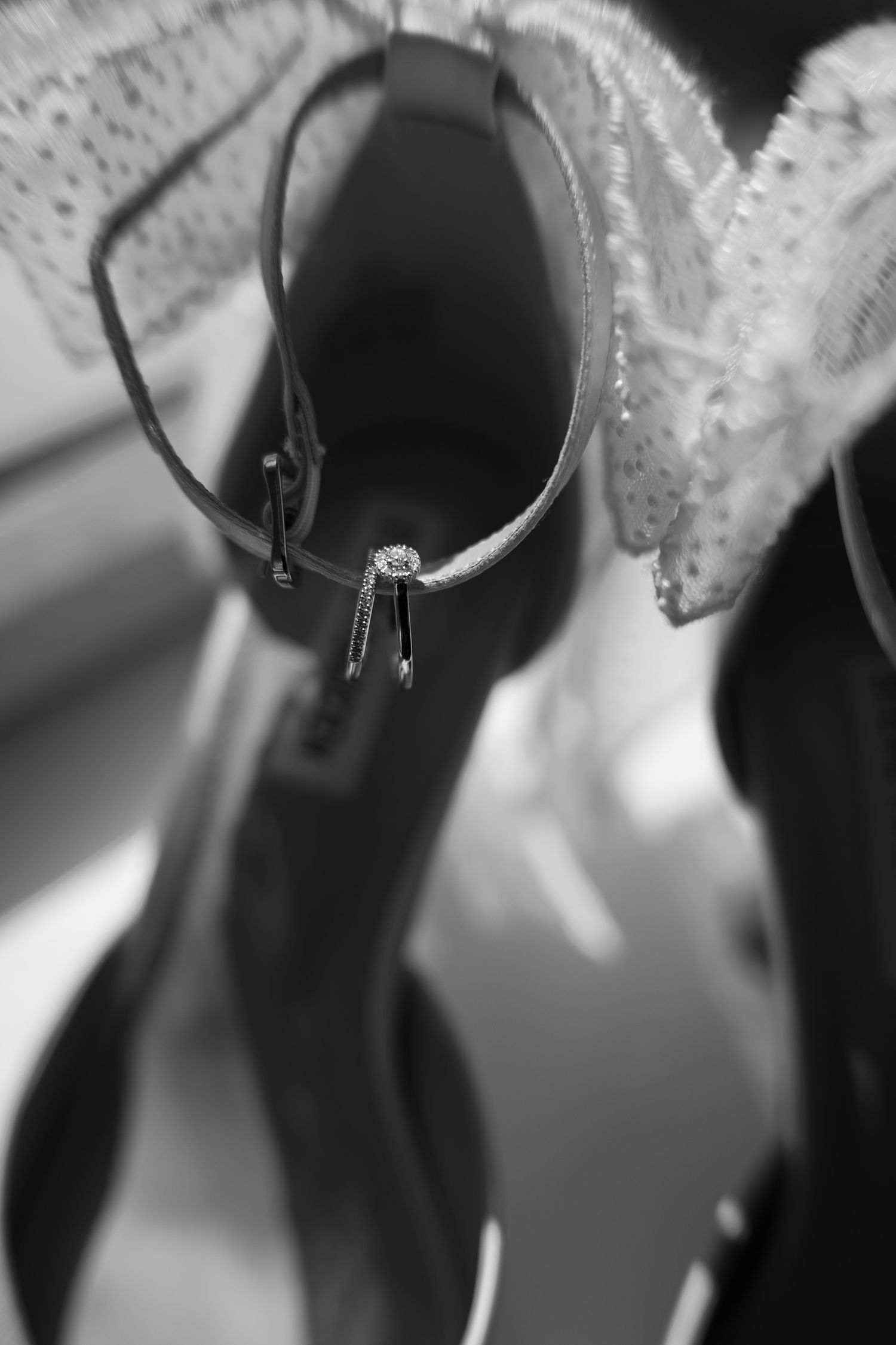Black and white photo of elegant high heel shoes with a sparkling diamond ring draped over them and lace fabric.