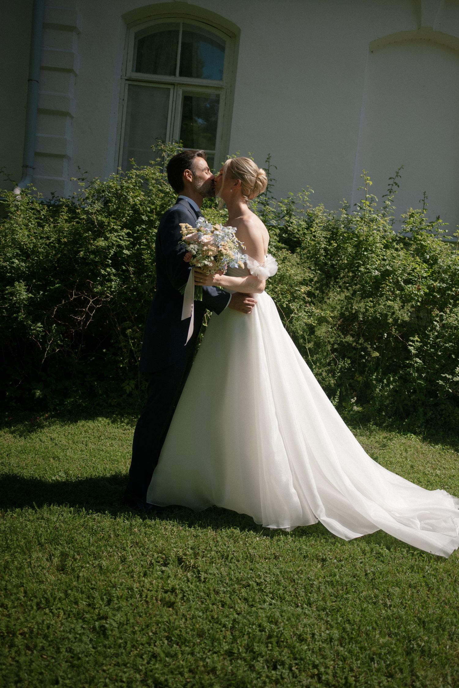 Bride and groom share a romantic moment in a white wedding dress and dark suit, standing on green grass near a building.